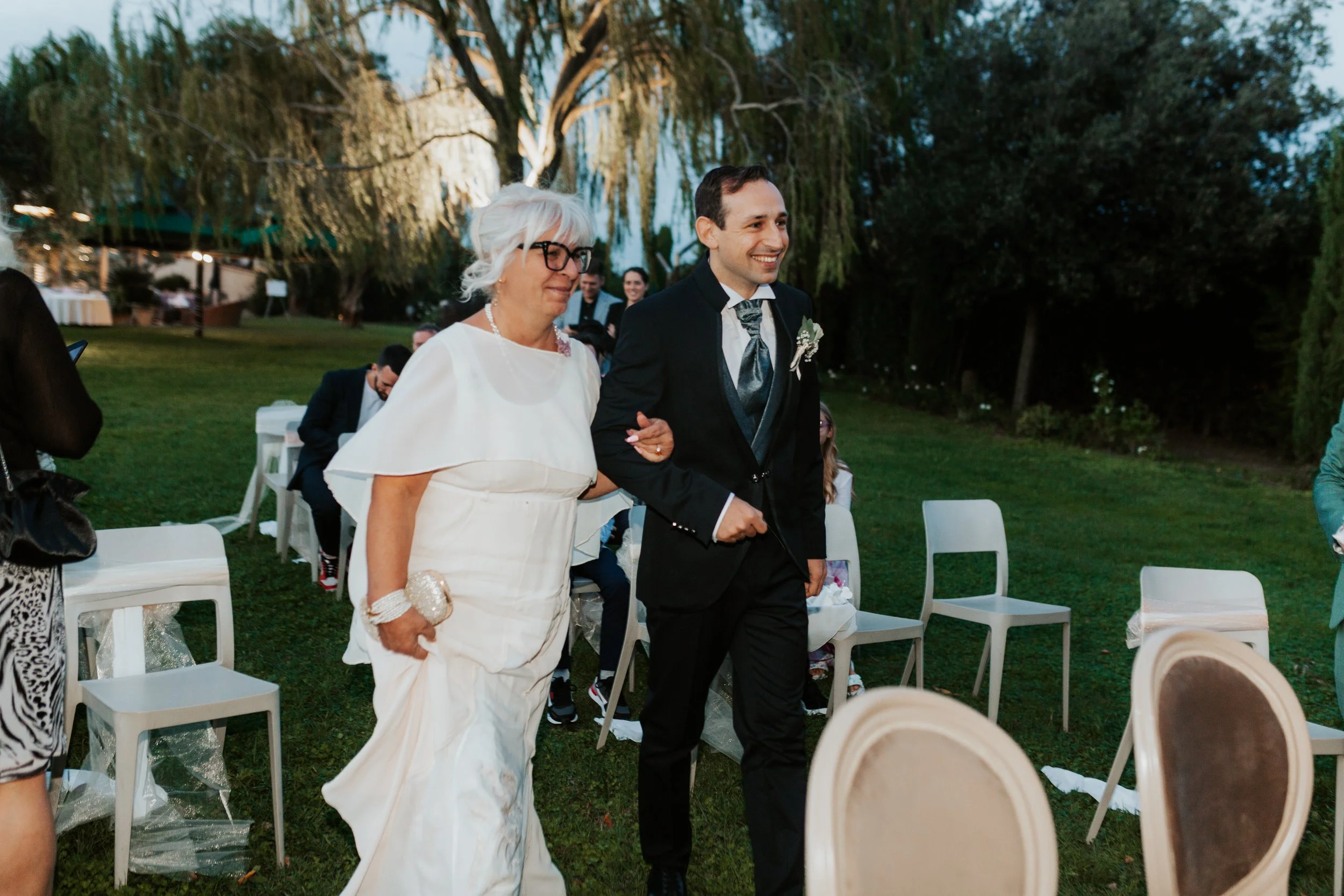 Una coppia di sposi cammina insieme all'aperto durante un matrimonio, circondati da sedie e alberi, con il cielo serale.