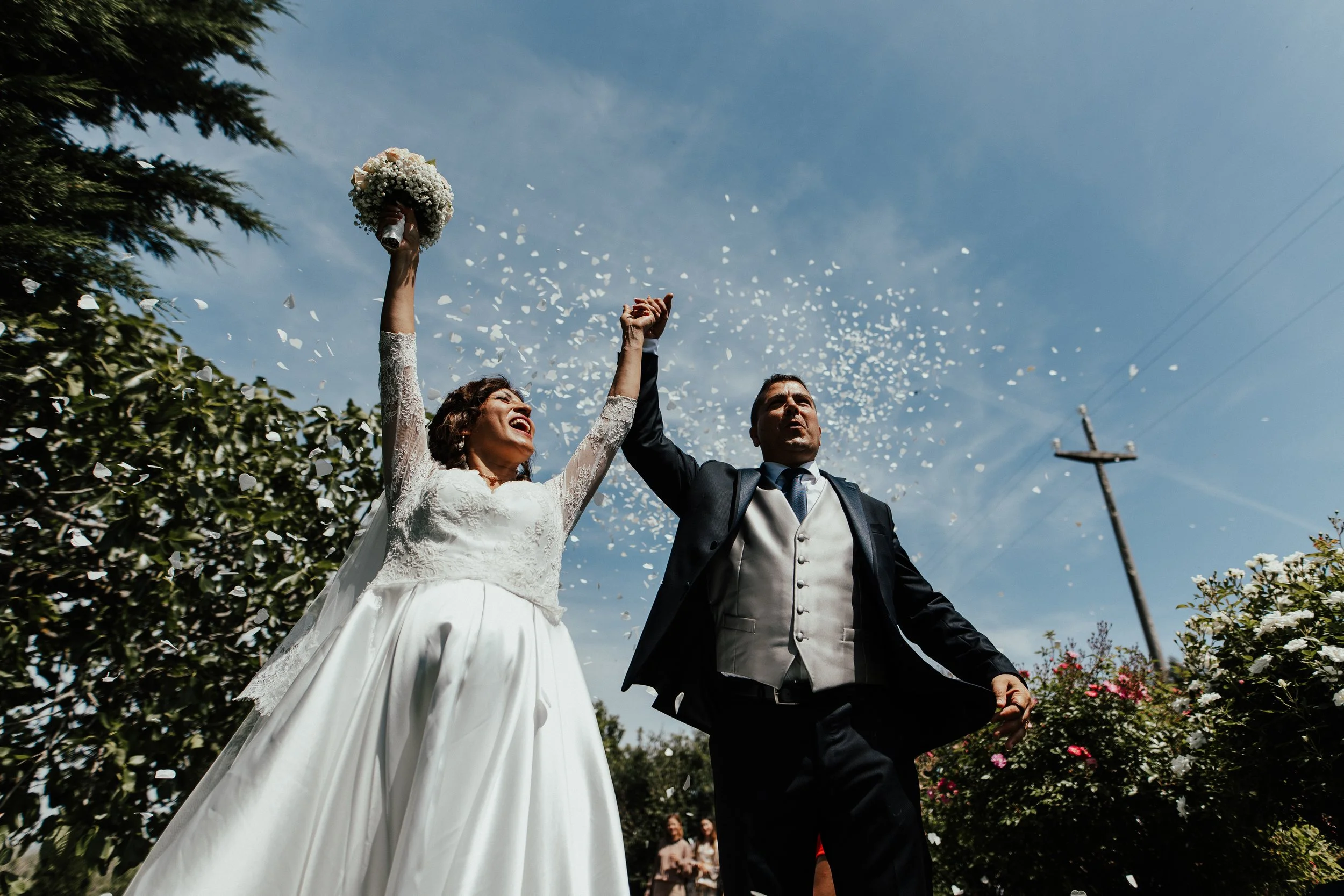 Sposi felici con mani alzate, stanno festeggiando sotto un cielo azzurro, circondati da fiori, durante il loro matrimonio.