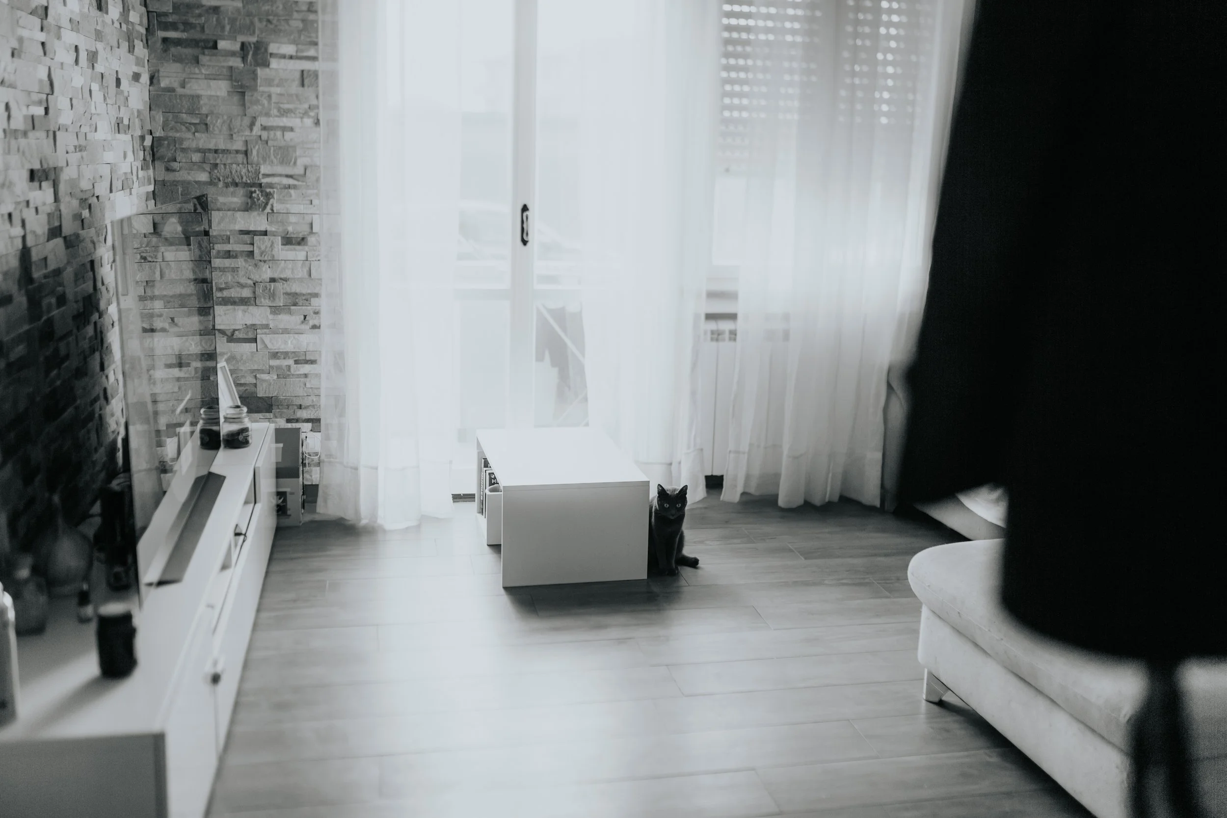 Interno di un soggiorno con parete in pietra, una TV, tende bianche, un gatto nero seduto vicino a una piccola tavola, e un divano chiaro.