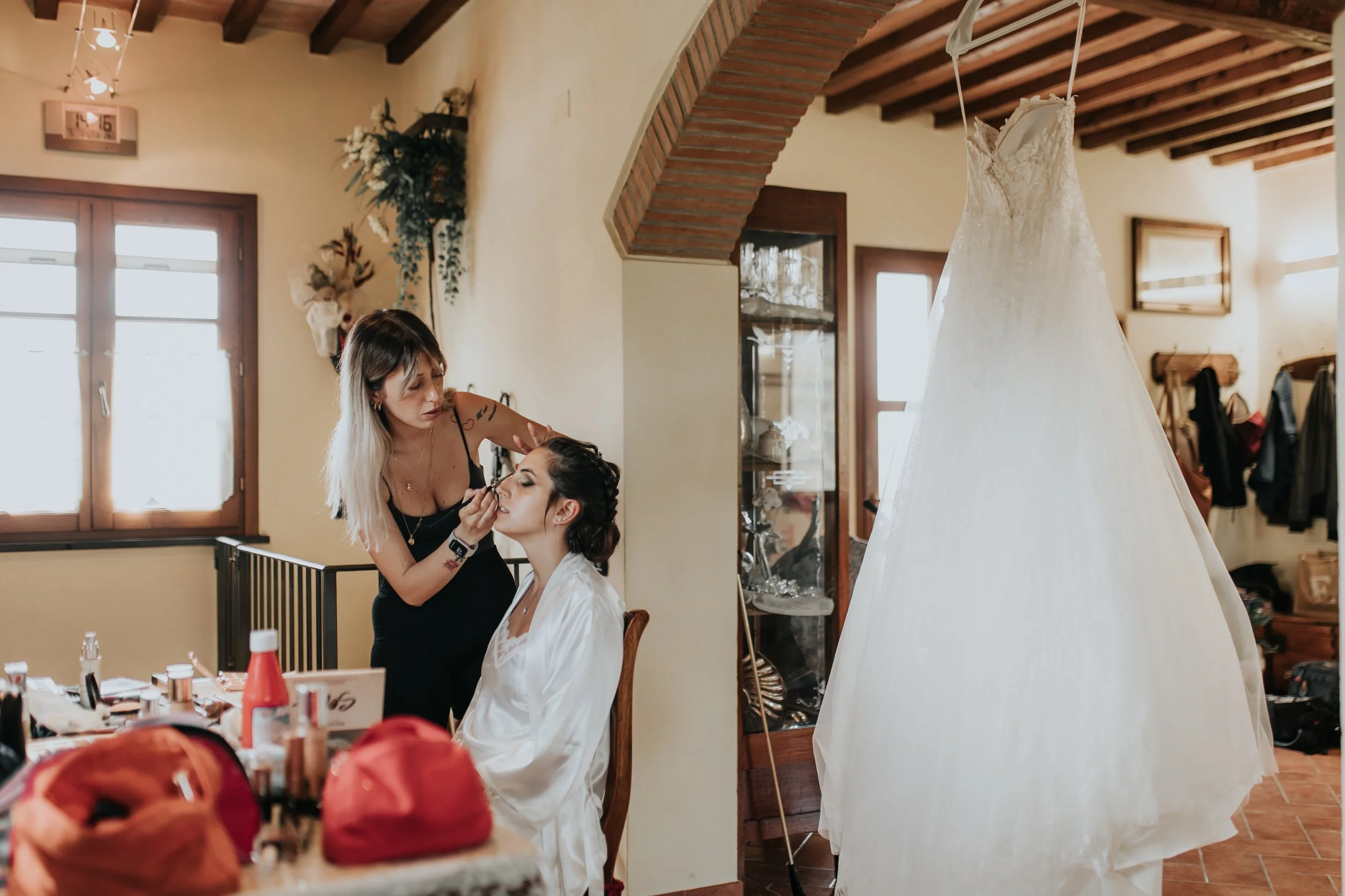 Una donna che fa il trucco a un'altra donna con un abito da sposa, in una stanza domestica, con una gonna di matrimonio appesa a un gancio.