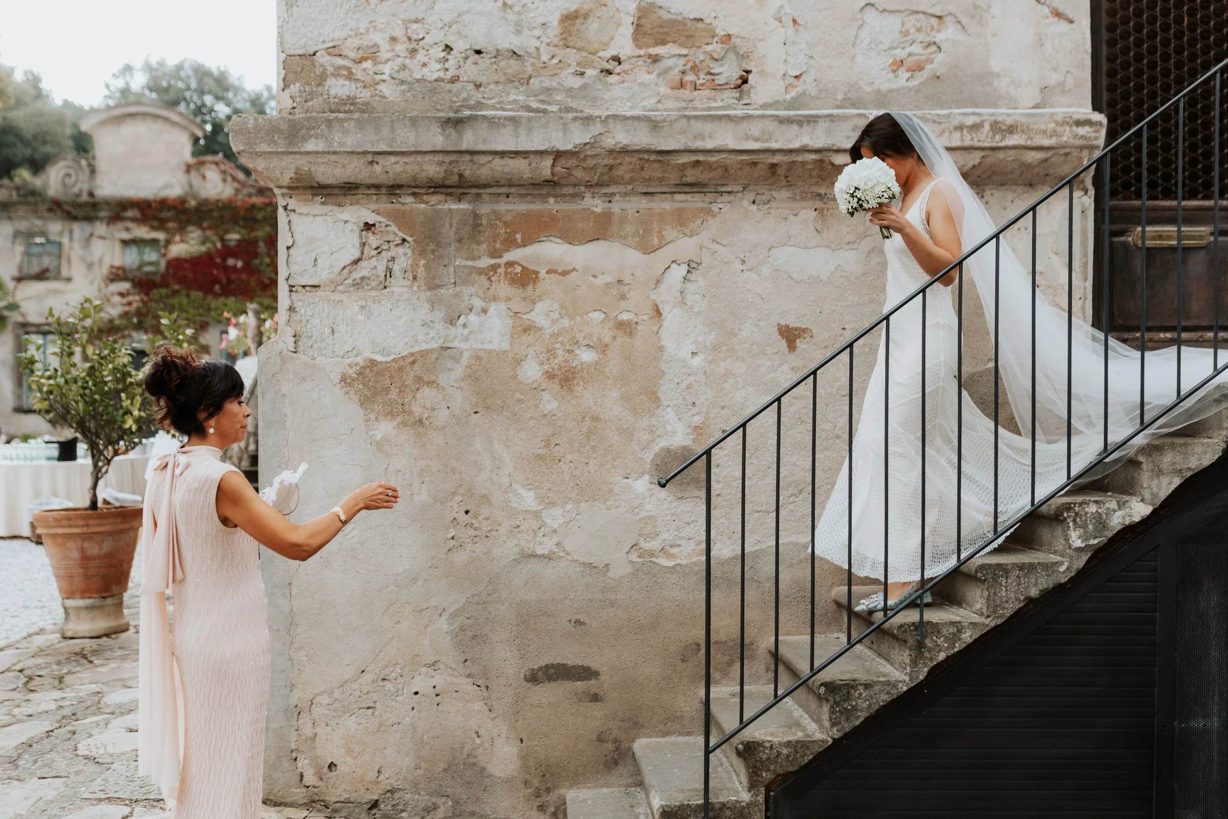 Una sposa con abito bianco e velo discende le scale mentre riceve un regalo da una donna con abito rosa chiaro, sullo sfondo un muro antico.