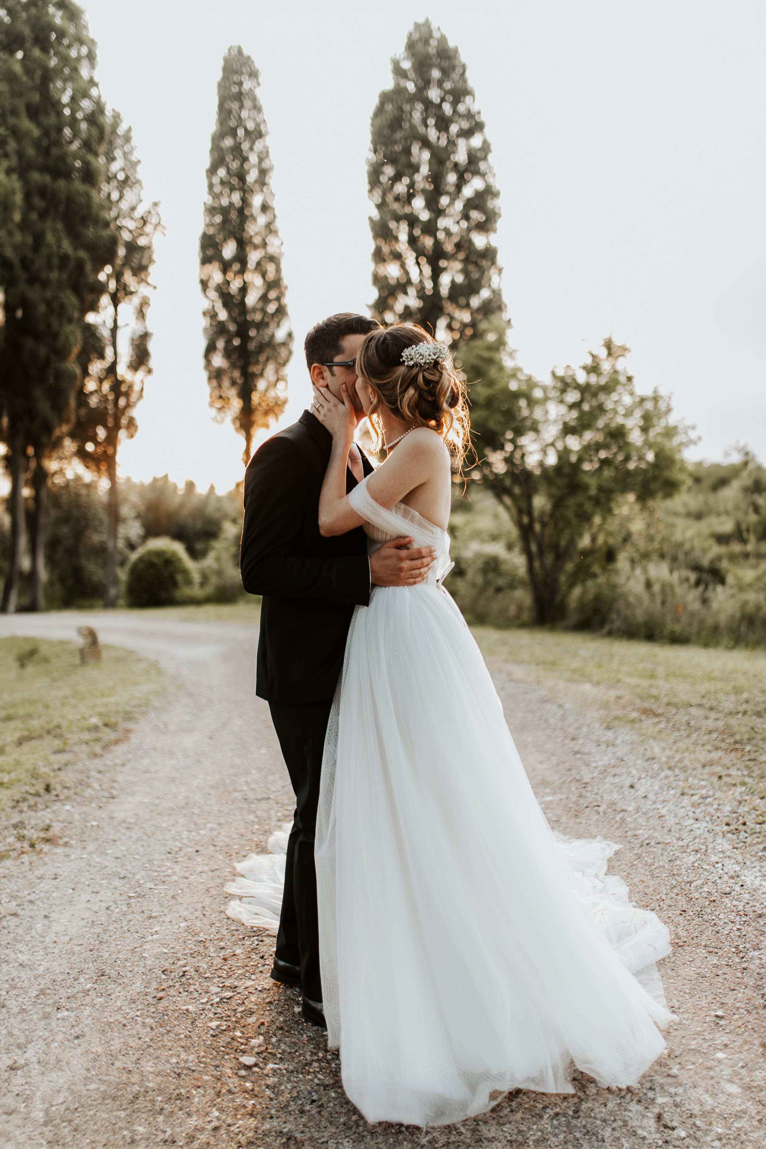 Una coppia di sposi si bacia all'aperto durante il tramonto, con alberi al tramonto sullo sfondo.