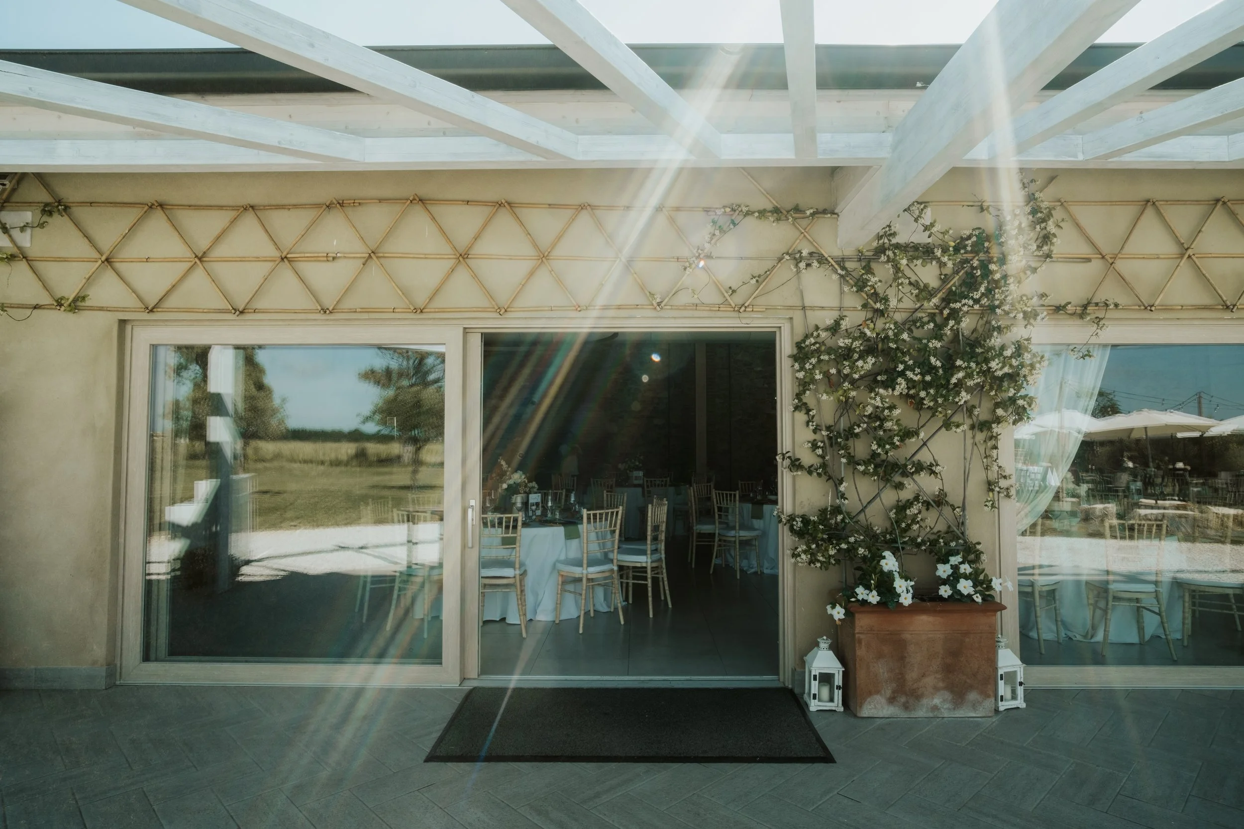 Entrata di una sala da pranzo decorata con piante e luci solari, con sedie di legno e tavoli rivestiti di tessuto bianco, vista esterna su un campo aperto.
