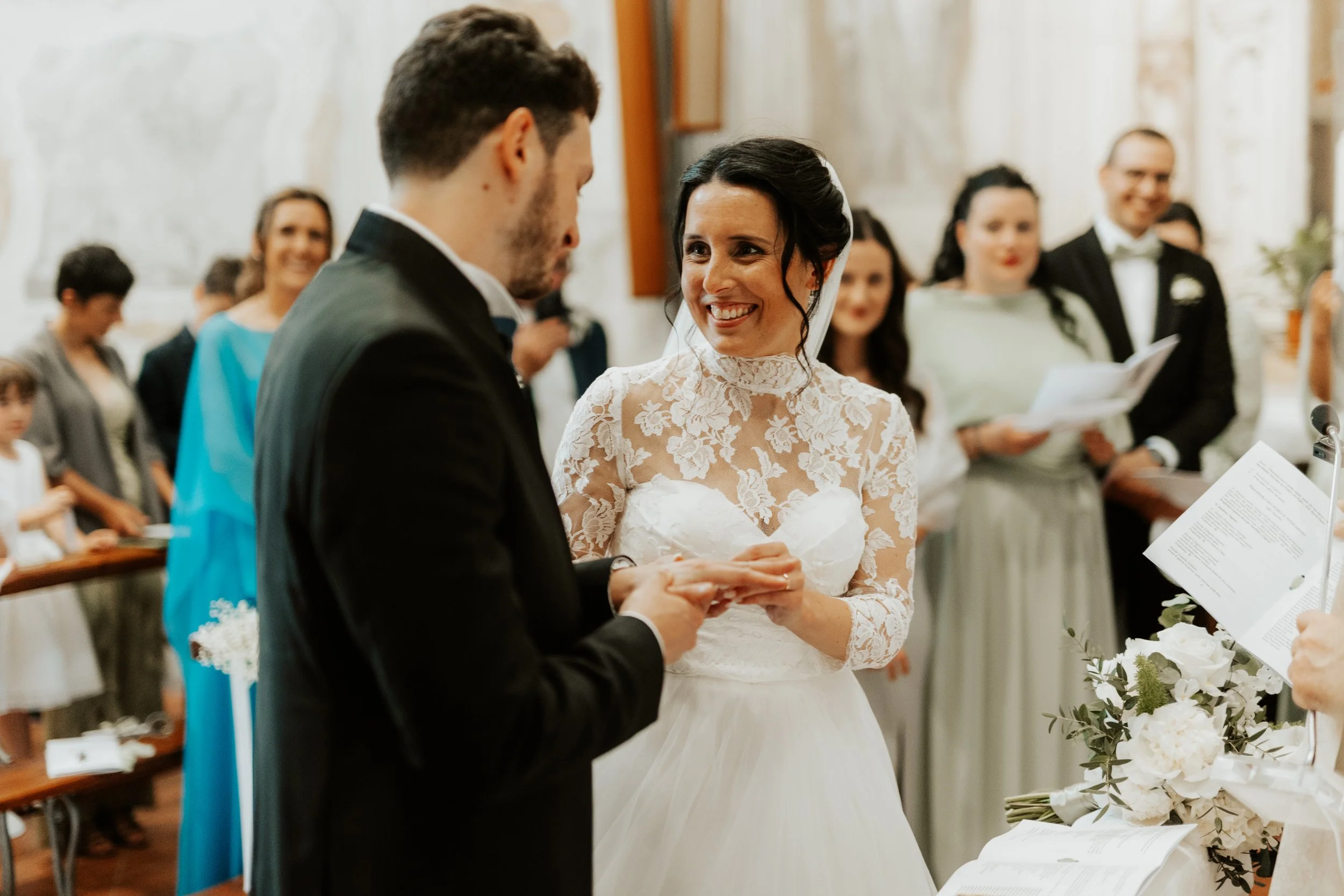 Una coppia di sposi si scambia le firme durante la cerimonia in chiesa, circondati da invitati sorridenti.