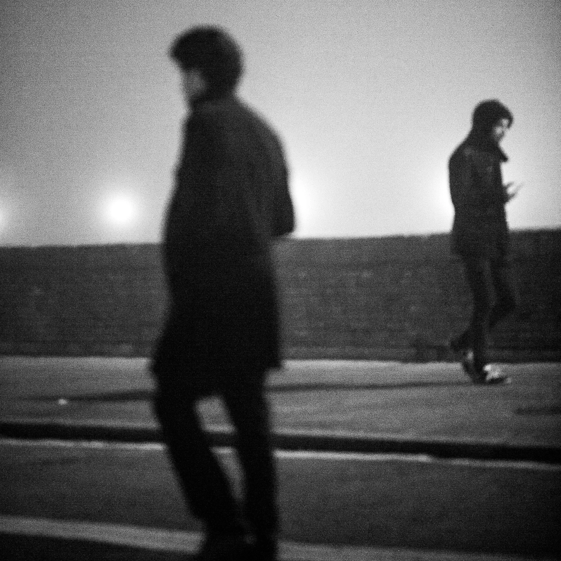 Due persone camminano al crepuscolo o di notte, una che guarda lo smartphone, in una scena in bianco e nero, in un'area all'aperto con una recinzione o muro di fondo.