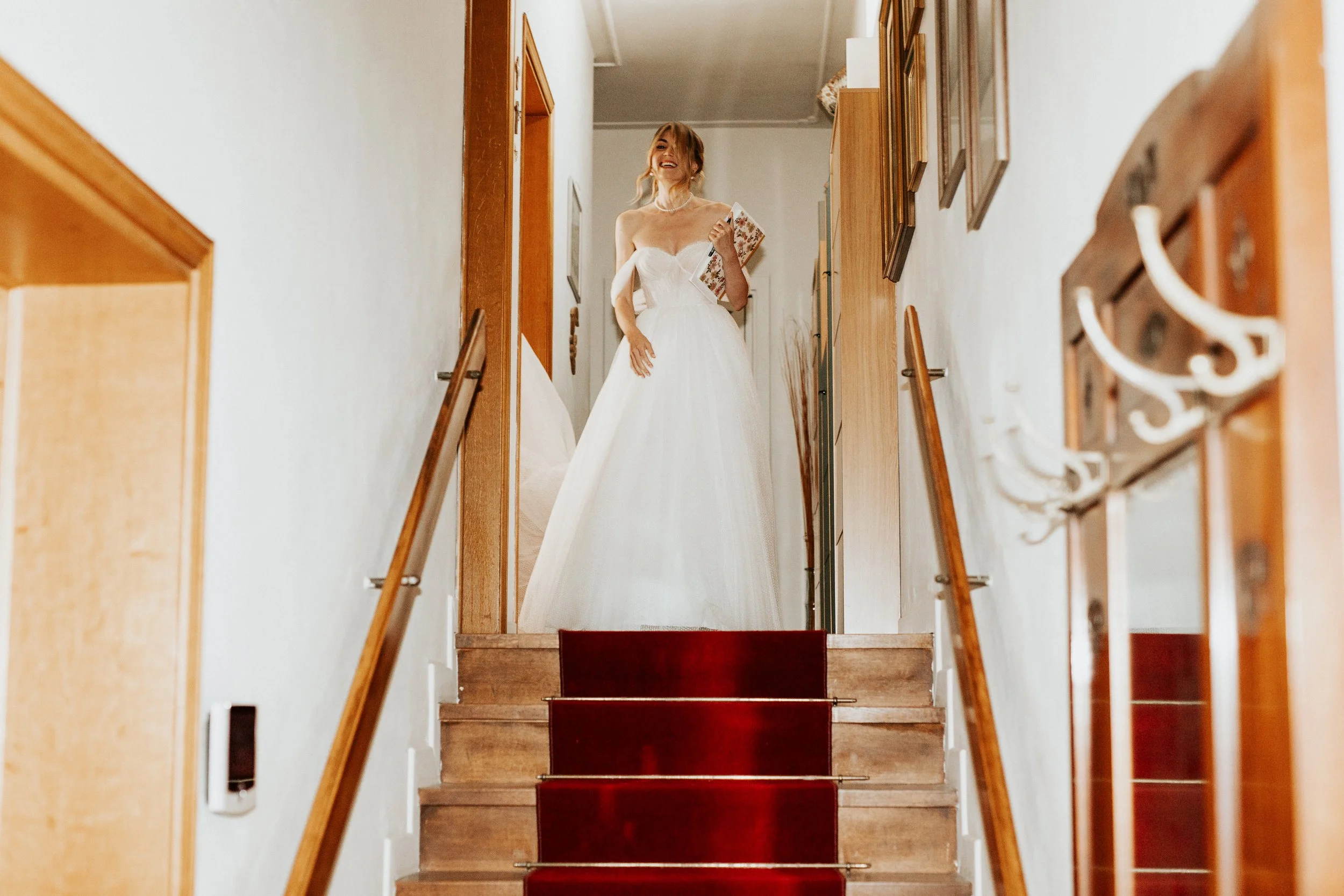 Una donna vestita con un abito da sposa bianco che sorride in una casa, vista dal basso delle scale
