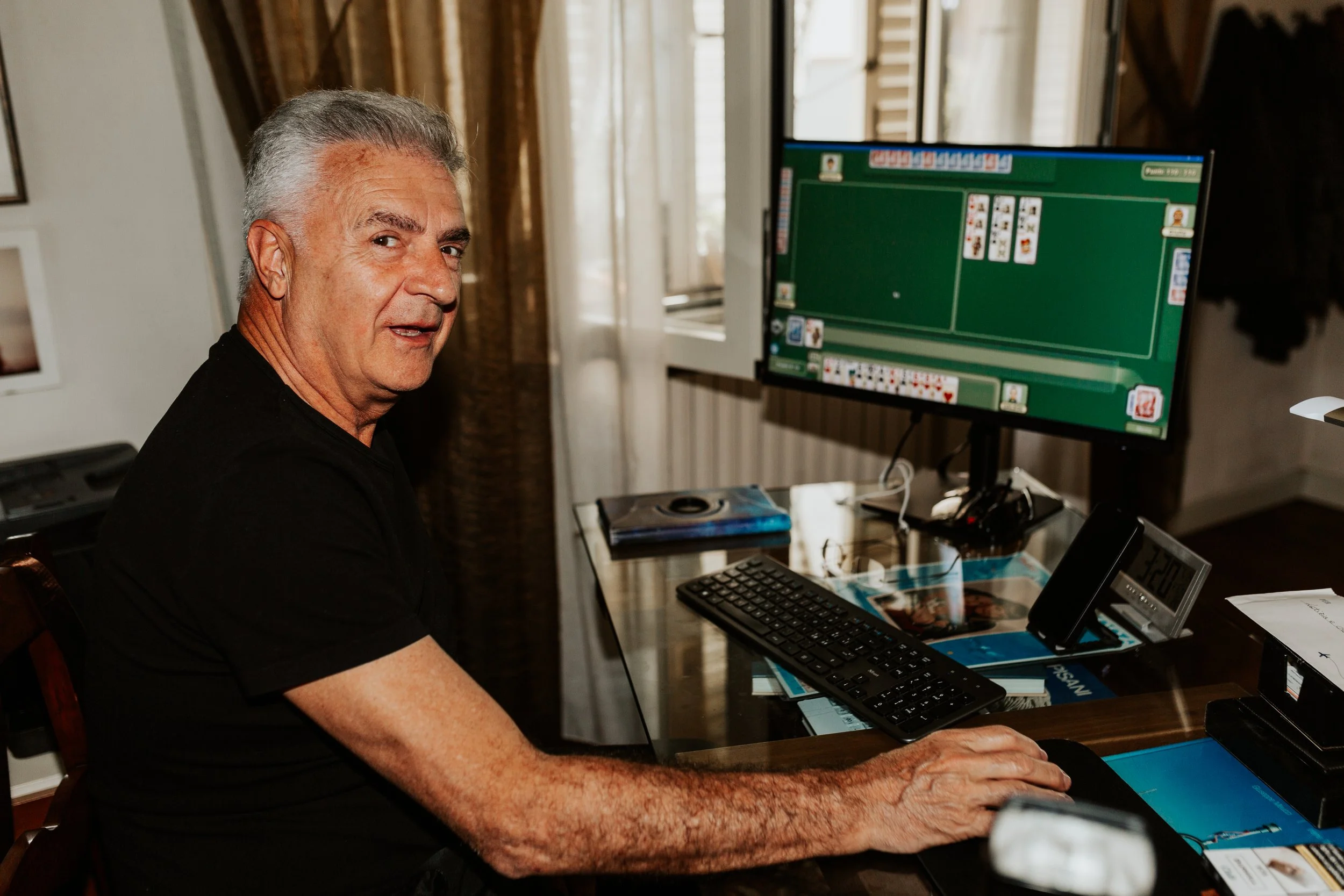 Uomo anziano che gioca a carte sul computer a casa, con una schermata di poker online visibile sul monitor.