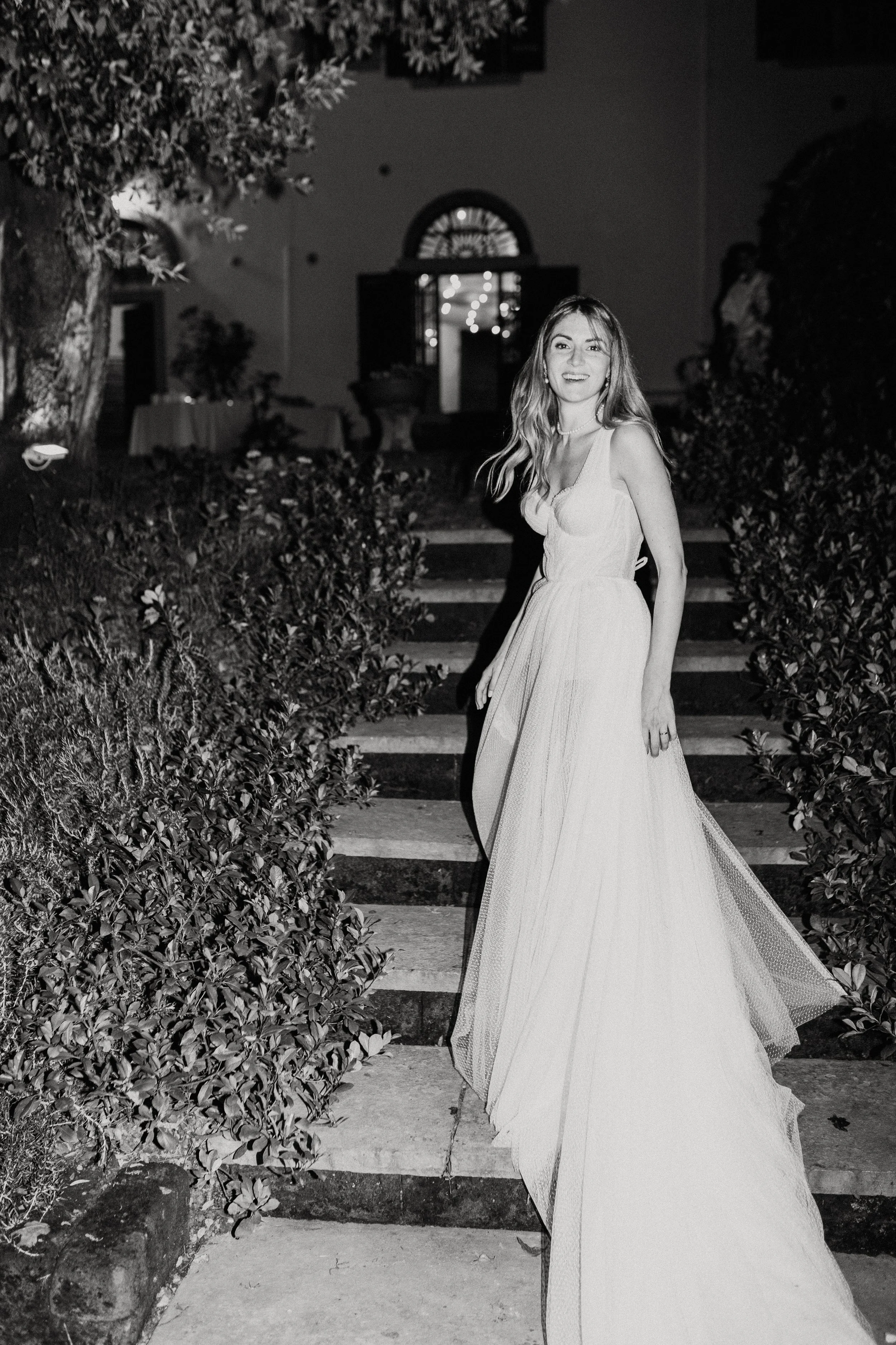 Una donna con un lungo abito bianco cammina lungo una scalinata di pietra, circondata da cespugli e alberi, di notte.