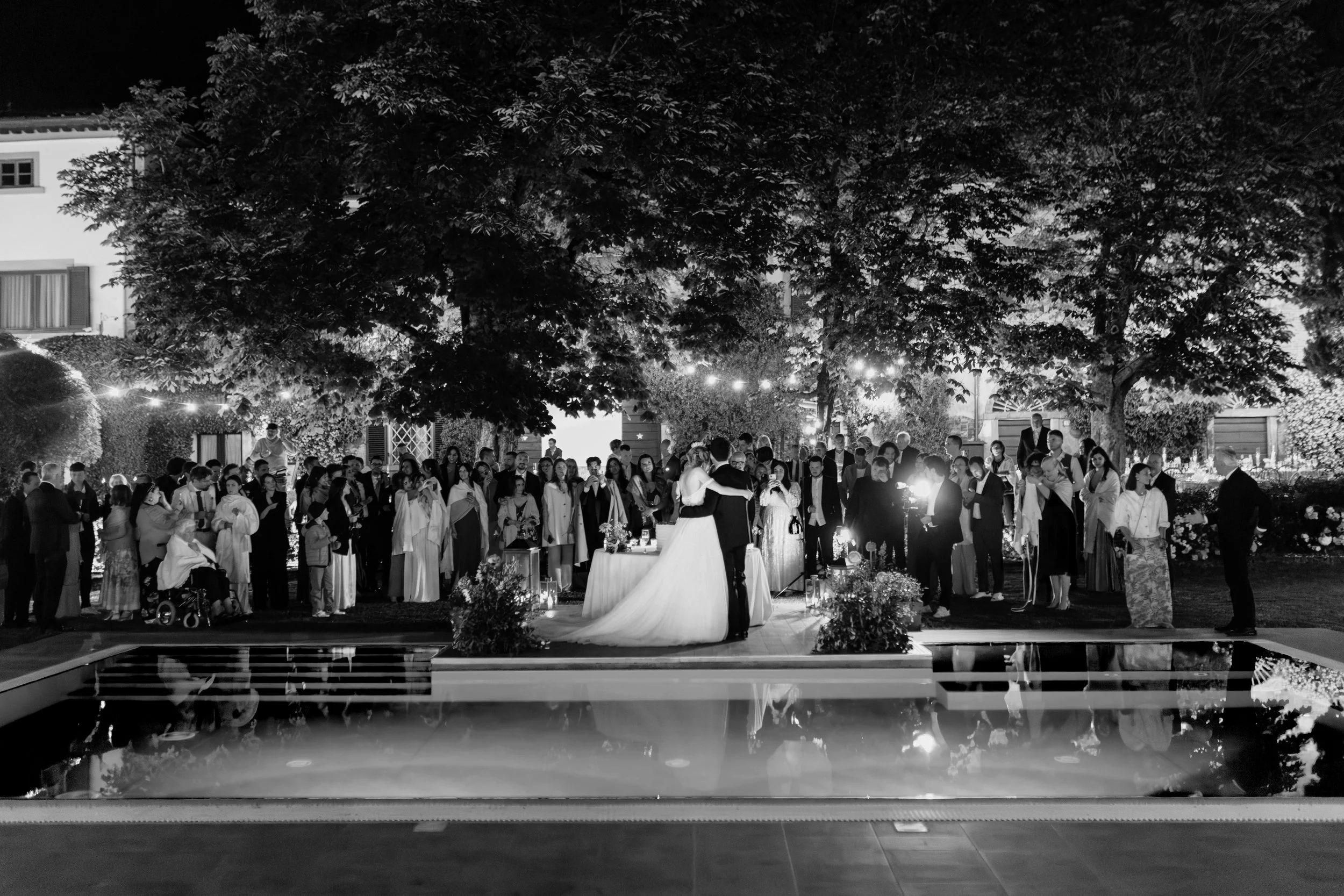 Coppia di sposi che si abbracciano al centro di una festa di matrimonio all'aperto, con molti ospiti, durante la sera, intorno a un'area con alberi e luci decorative, riflessa in una piscina.