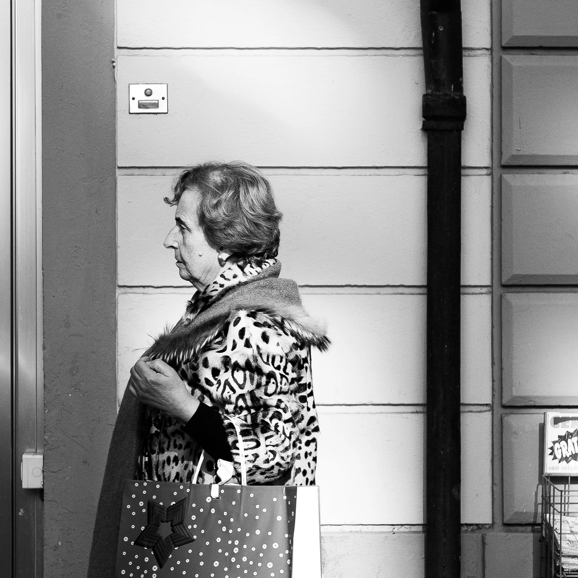 Una donna anziana con abito leopardato e sciarpa, che tiene una borsa decorata con pallini, guarda decisa a destra in un contesto urbano in bianco e nero.