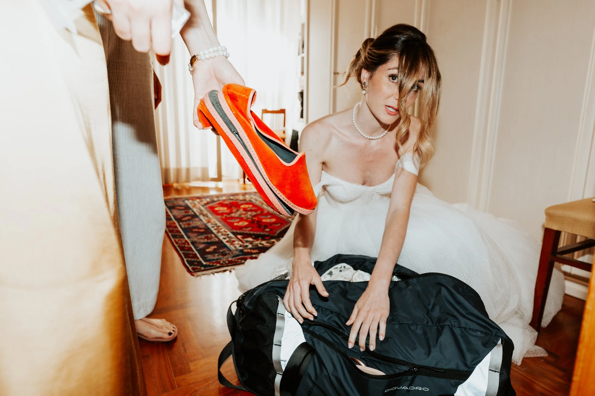 Una donna in abito bianco seduta su un letto, mentre riceve una scarpa da un'altra persona