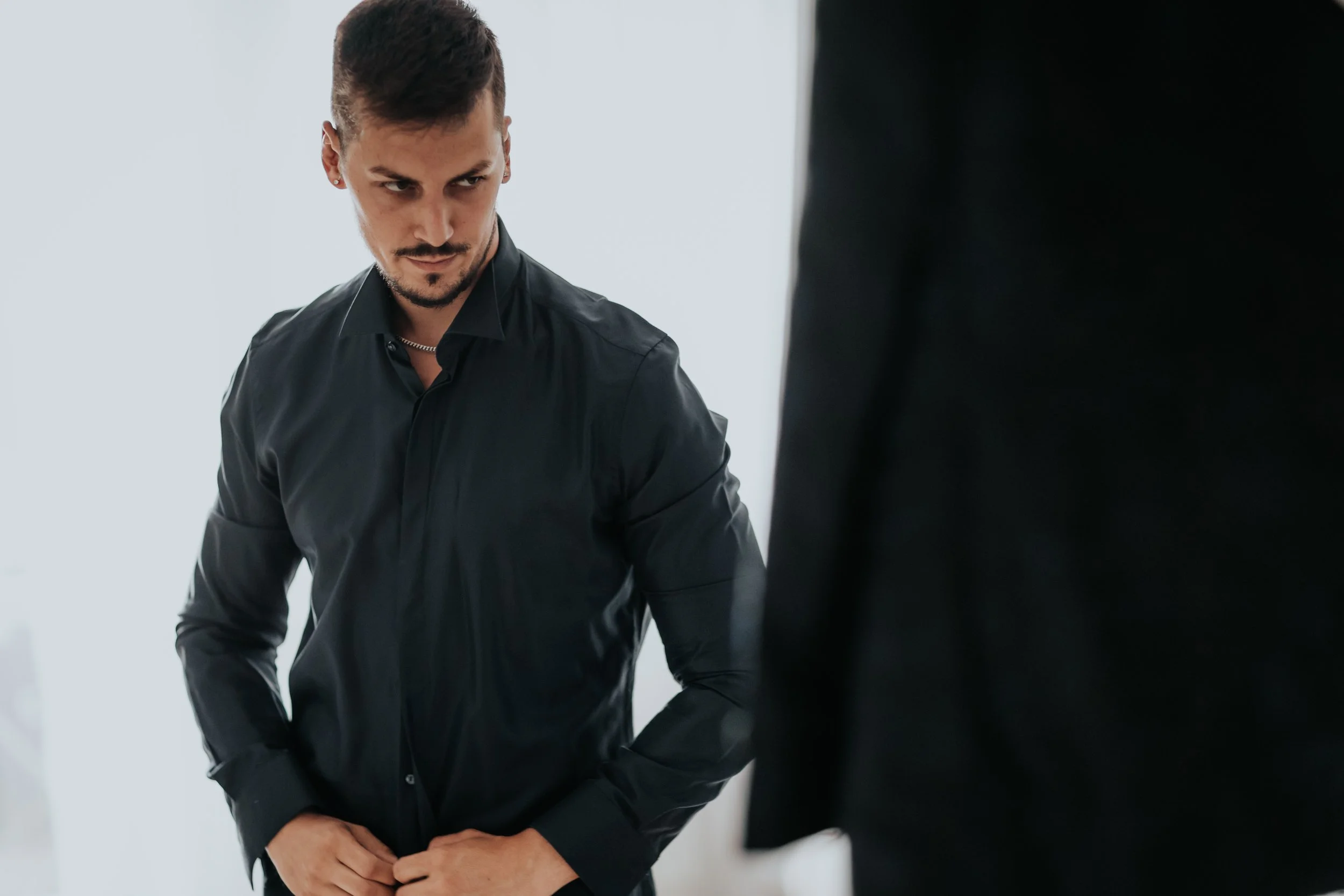 Uomo con capelli corti e barba che indossa una camicia nera con le mani sui bottoni, che guarda intensamente a destra
