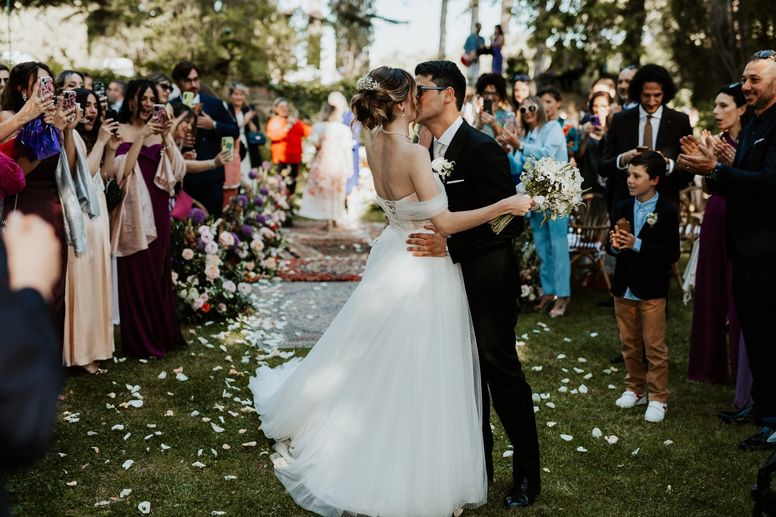 Coppia di sposi che si bacia durante il matrimonio, circondata dagli invitati che scattano foto e applaudono all'aperto. Villa dal Pozzo
