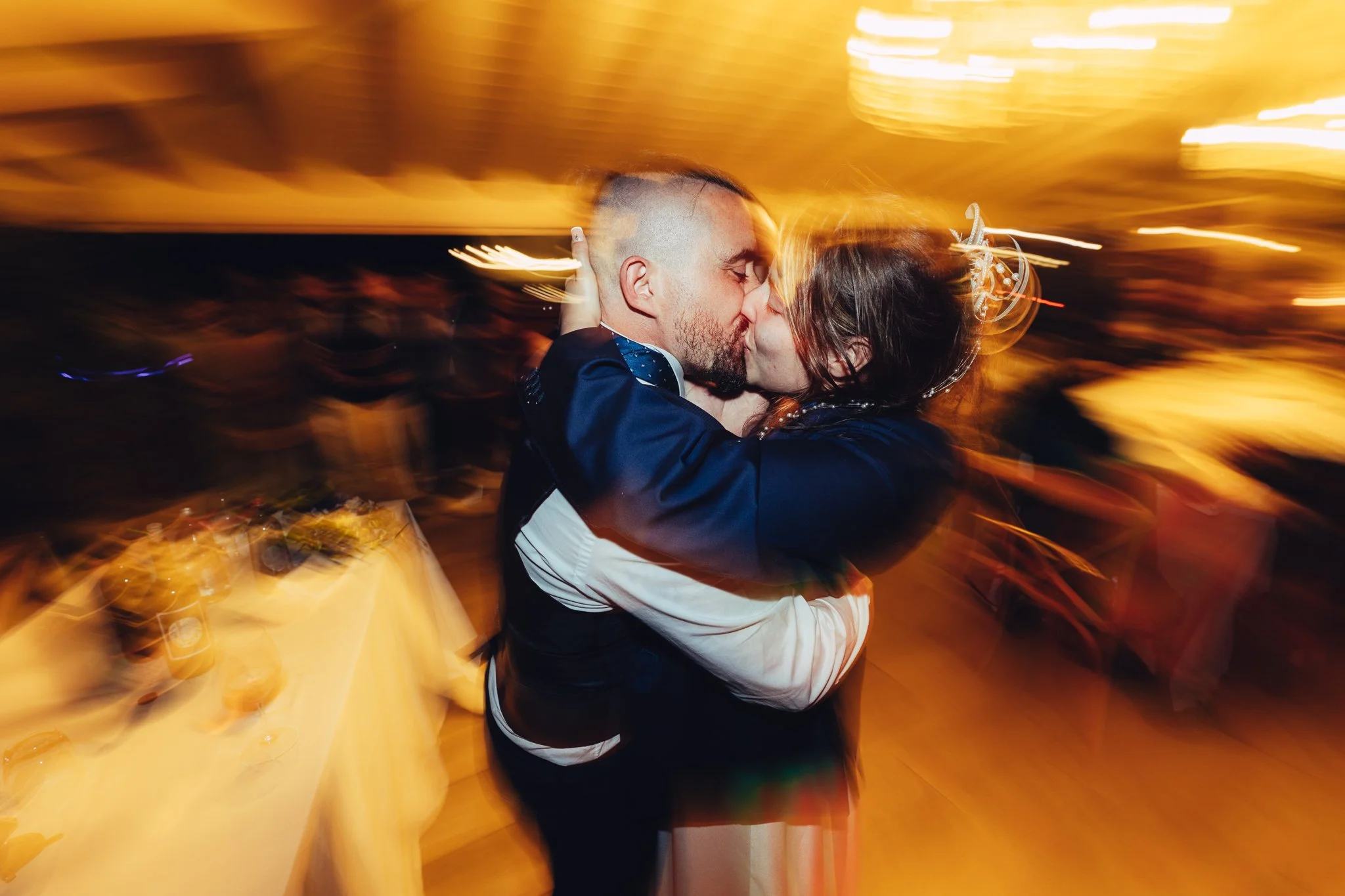 Due persone si baciano durante un ballo, con luci calde e movimento sfocato sullo sfondo.