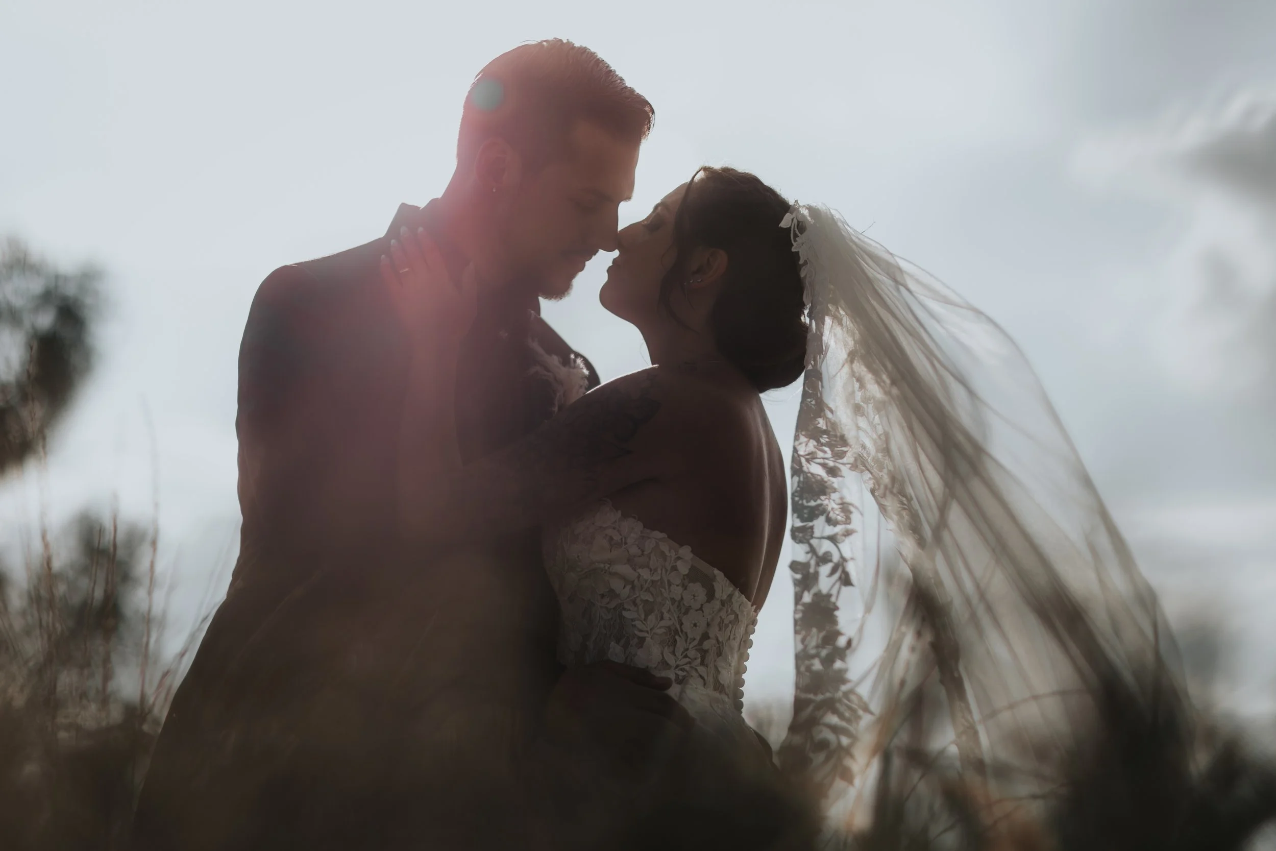 Una coppia di sposi si abbraccia e si avvicina in un contesto naturale, con il cielo sullo sfondo. Lei indossa un abito da sposa con un velo, lui un abito scuro.