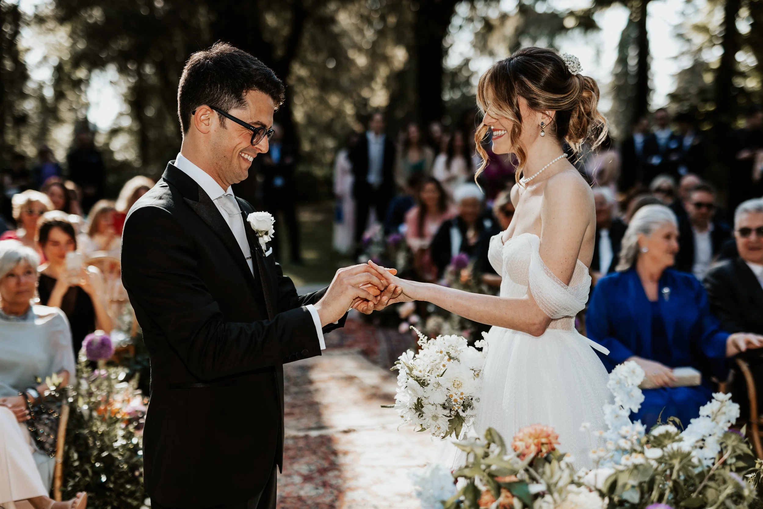 Una coppia di sposi durante il rito di matrimonio all'aperto, la donna in abito bianco e il uomo in completo nero, si scambiano le fedi tra gli applausi degli invitati.