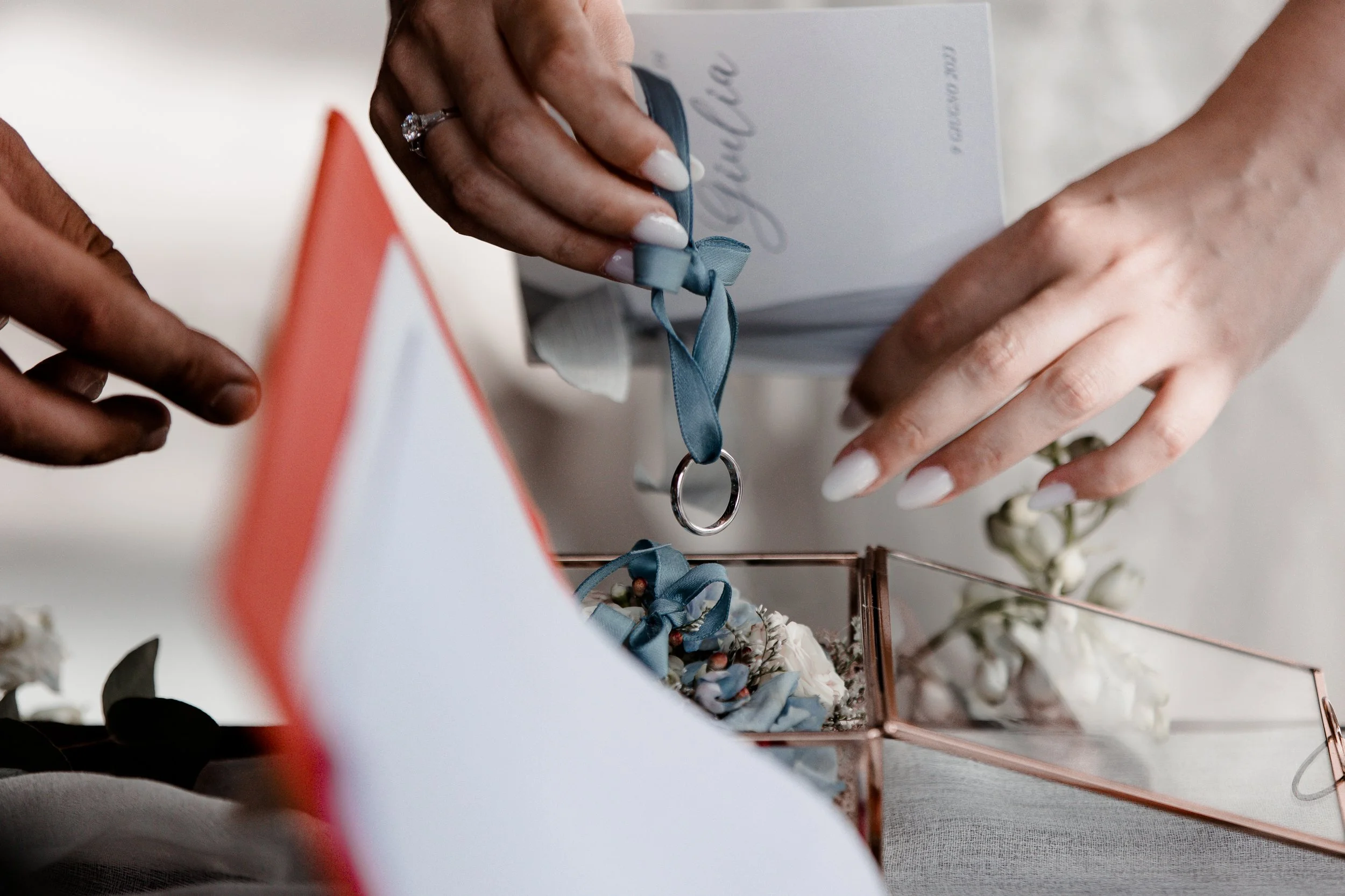 Due mani con un anello e unghie con smalto bianco, una che tiene una scatola di presentazione blu e l'altra che sistema una scatola con fiori bianchi e un anello in un contenitore di vetro con decorazioni blu.