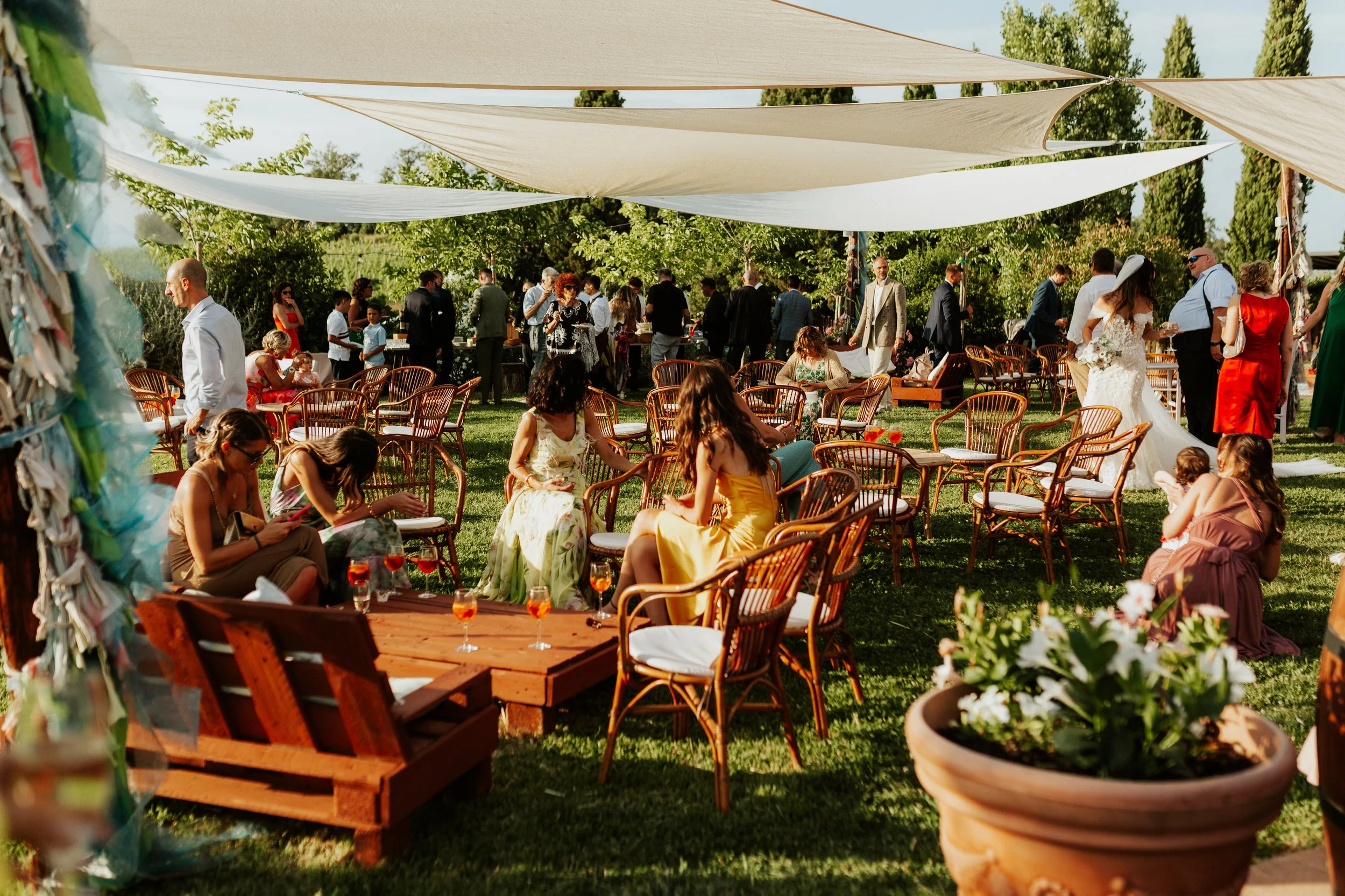 Un gruppo di persone festeggia un matrimonio in un giardino con sedie di legno e ombrelloni, alcune persone sono sedute mentre altre sono in piedi e socializzano. Agriturismo Cà Lo Spelli. Del Carlo Catering