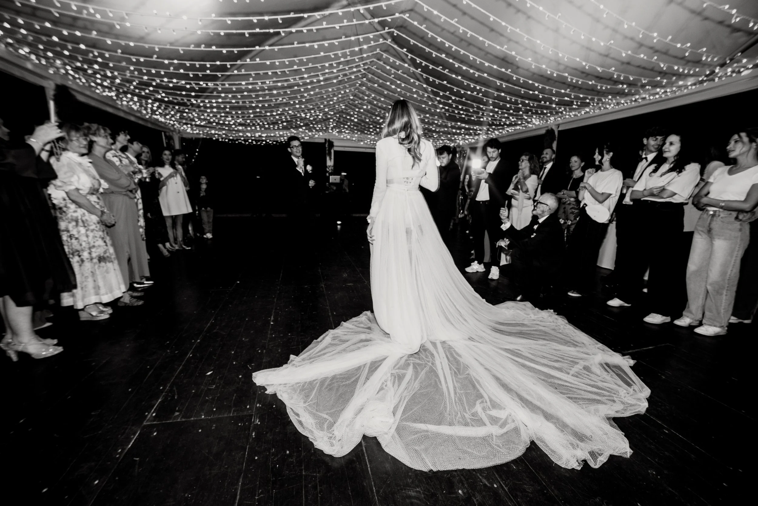 Una sposa in abito da matrimonio con gonna lunga e velo, circondata da amici e parenti durante una festa di matrimonio, sotto decorazioni di luci sul soffitto.
