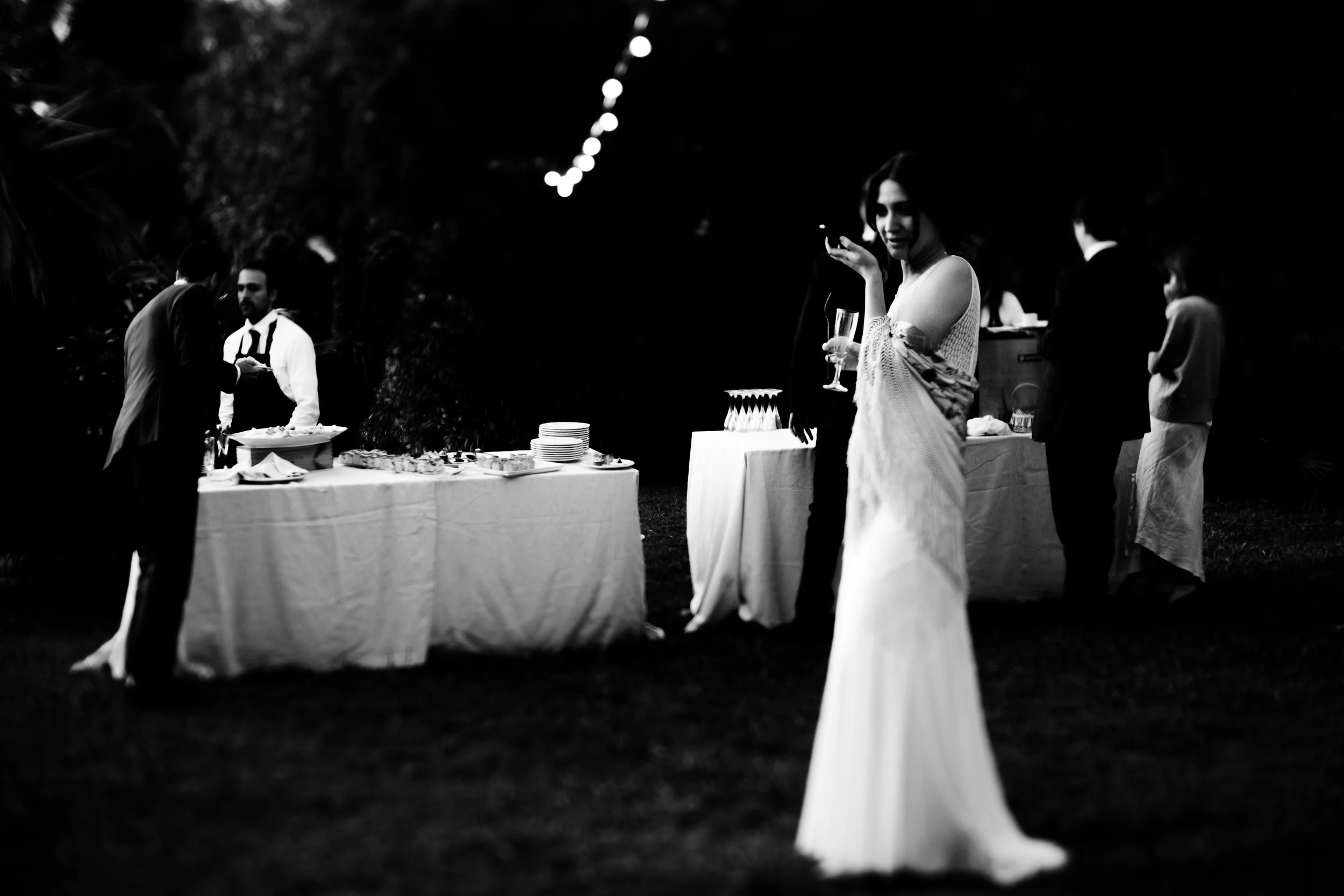 Scena di un ricevimento all'aperto di notte, con persone eleganti che si servono cibo e bevande, in un ambiente romantico e tranquillo.