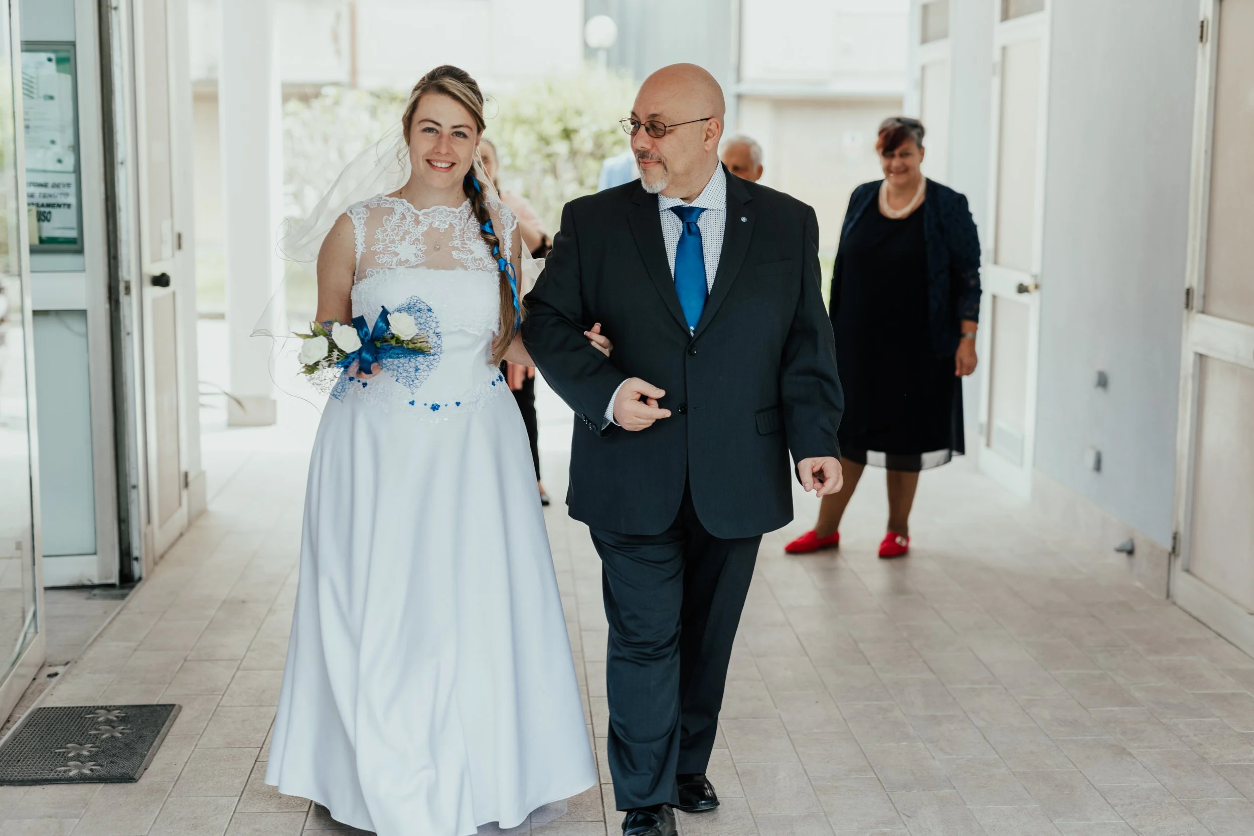 Una sposa e un uomo in abito elegante camminano insieme, circondati da altre persone, in un ingresso di un edificio, probabilmente in occasione di un matrimonio