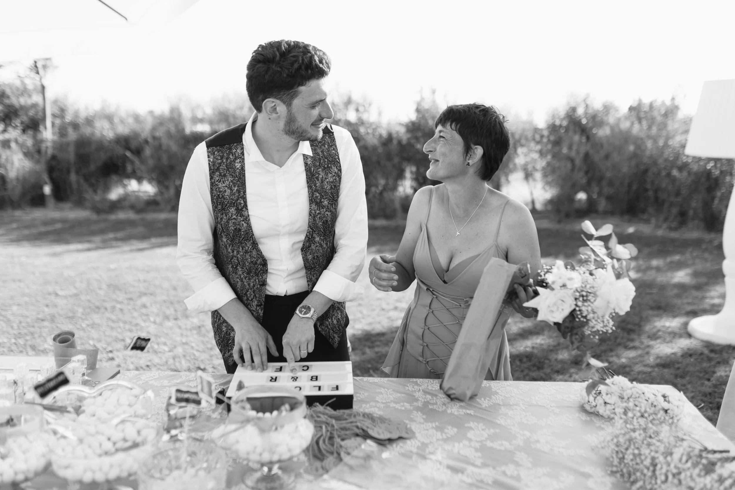 Un uomo e una donna sorridenti si scambiano uno sguardo mentre interagiscono in un evento all'aperto, con un tavolo decorato con fiori e dolci.