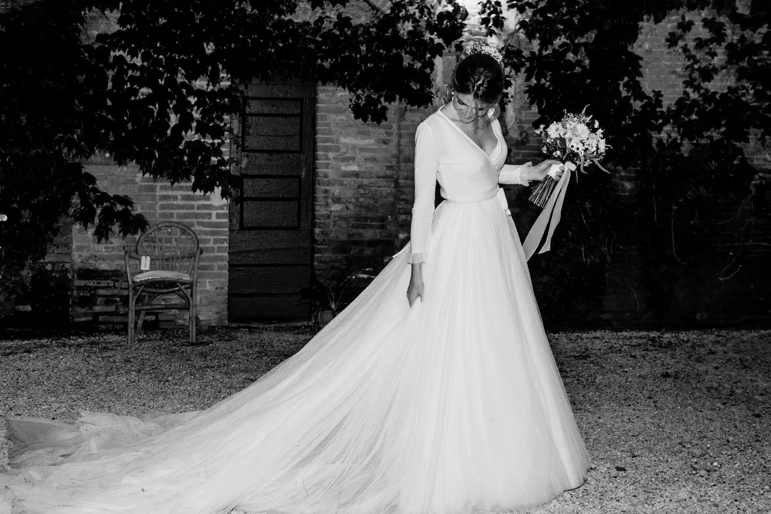 Una sposa con abito bianco lungo con una lunga coda, tiene un bouquet di fiori, si trova all'aperto di sera, con un muro di mattoni e un'area con alberi sullo sfondo.