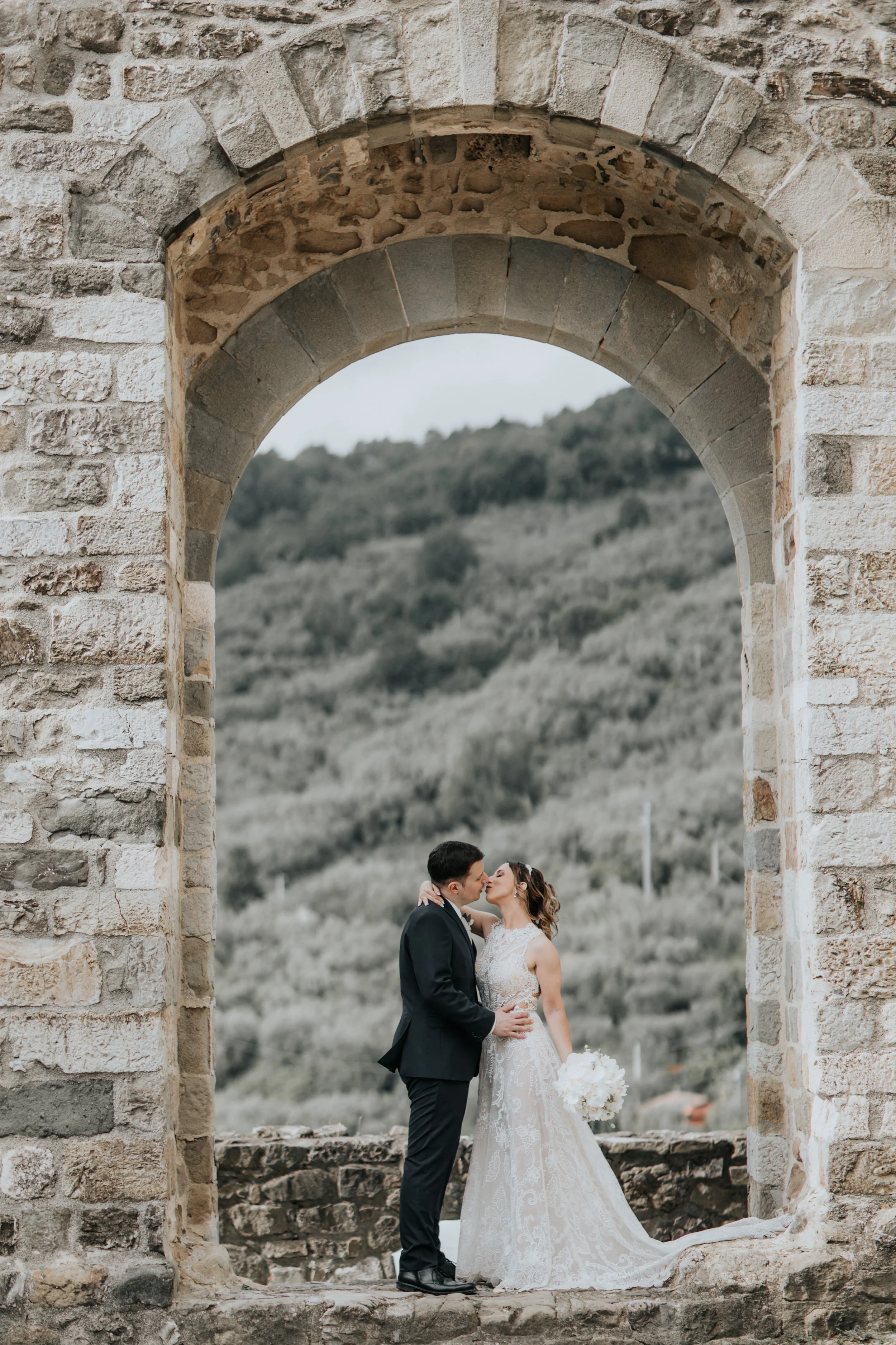 Una coppia di sposi si bacia sotto una vecchia arcata di pietra, con uno sfondo di colline verdi.