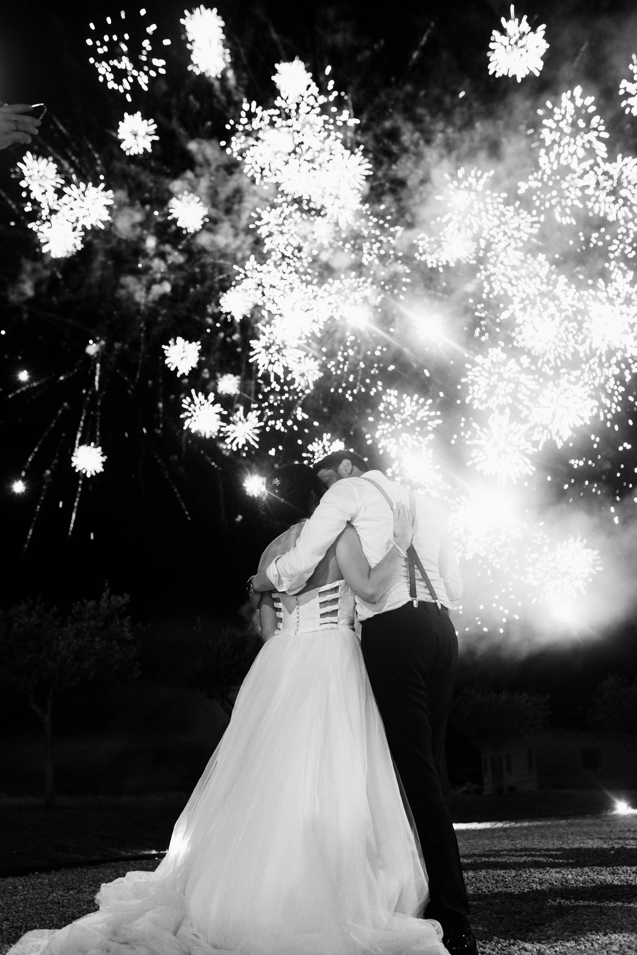 Due persone che si abbracciano sotto fuochi d'artificio durante una celebrazione notturna, una donna in abito bianco e un uomo in camicia e pantaloni neri.