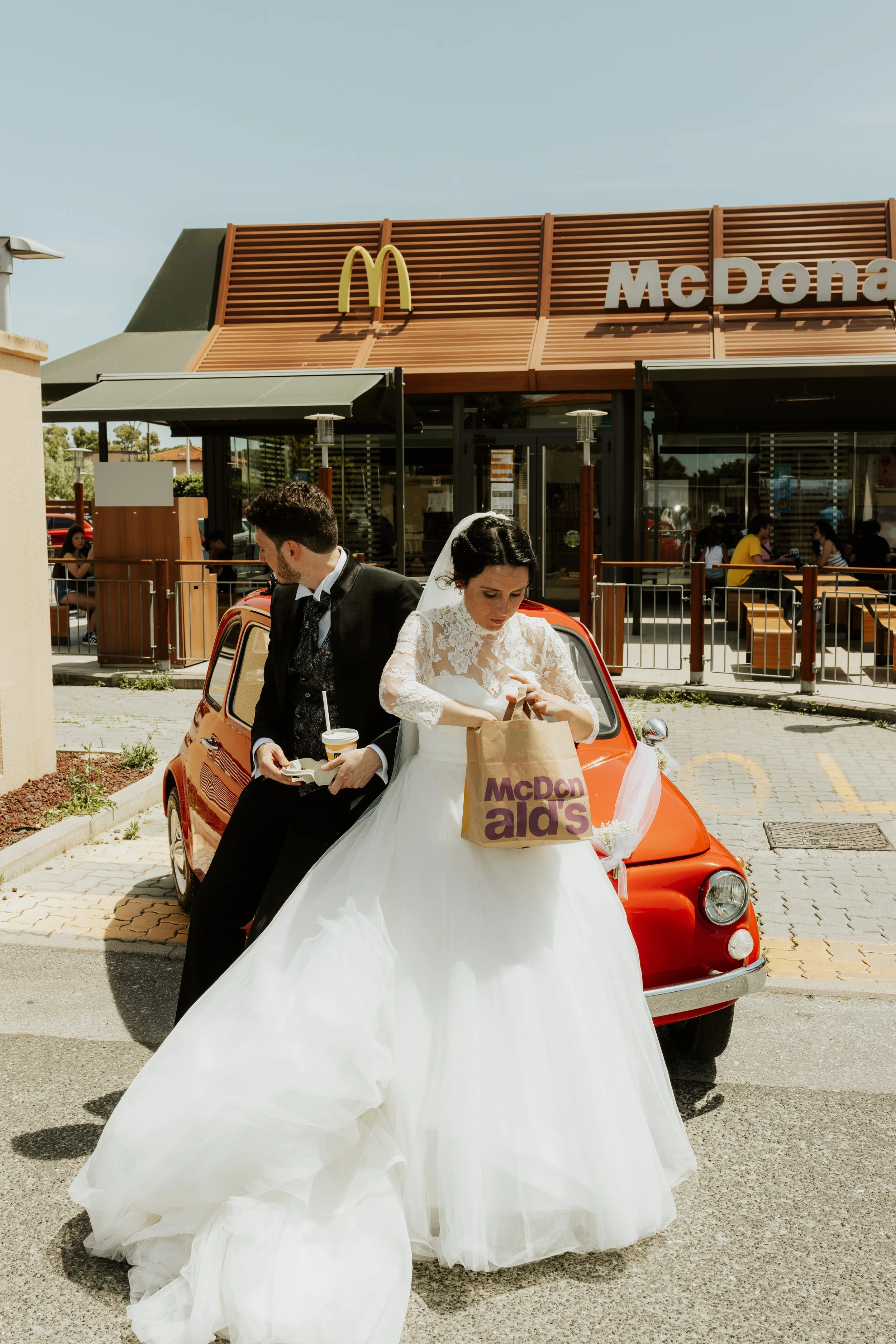 Sposi in abiti da matrimonio vicino a una macchina rossa davanti a un ristorante McDonald's.