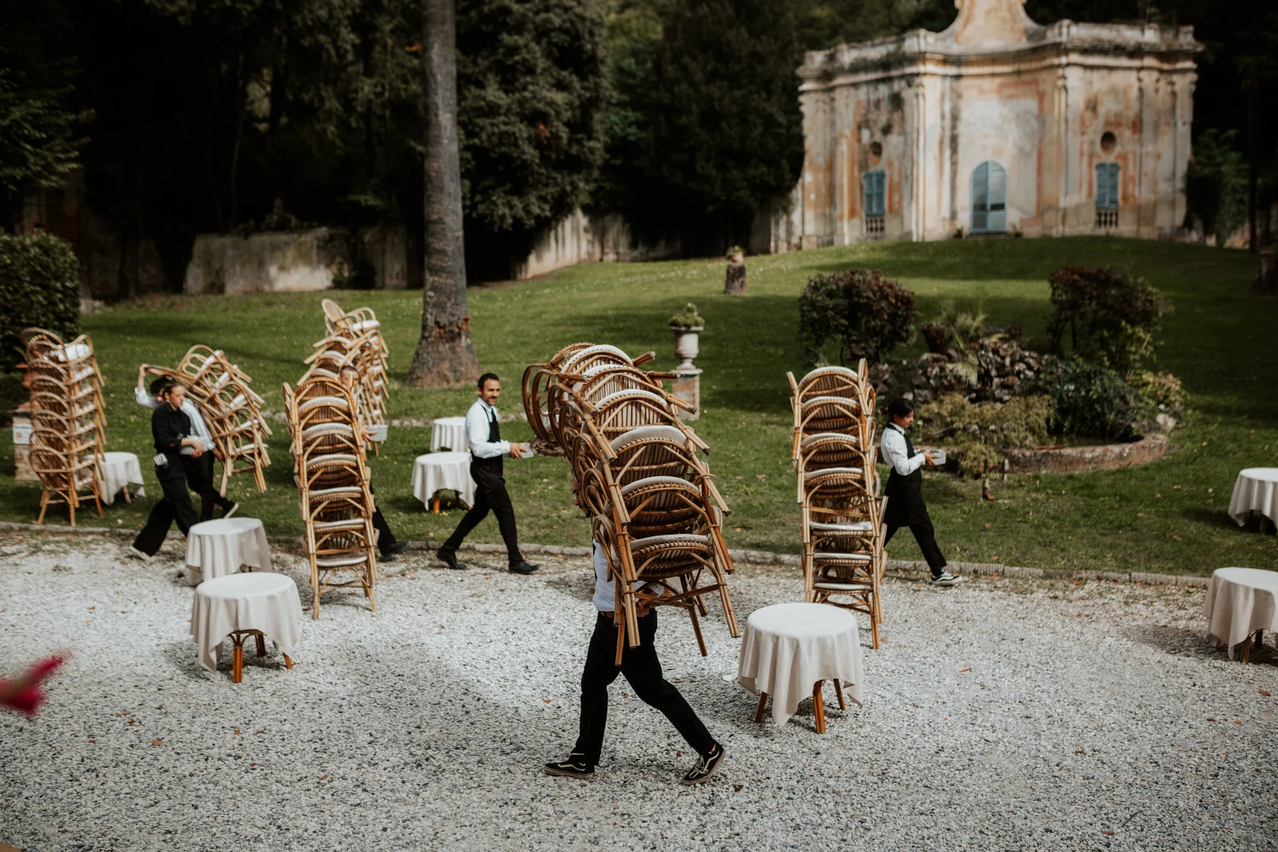 Camerieri trasportano sedie a un evento all'aperto in un giardino con un edificio storico sullo sfondo. Villa di Corliano
