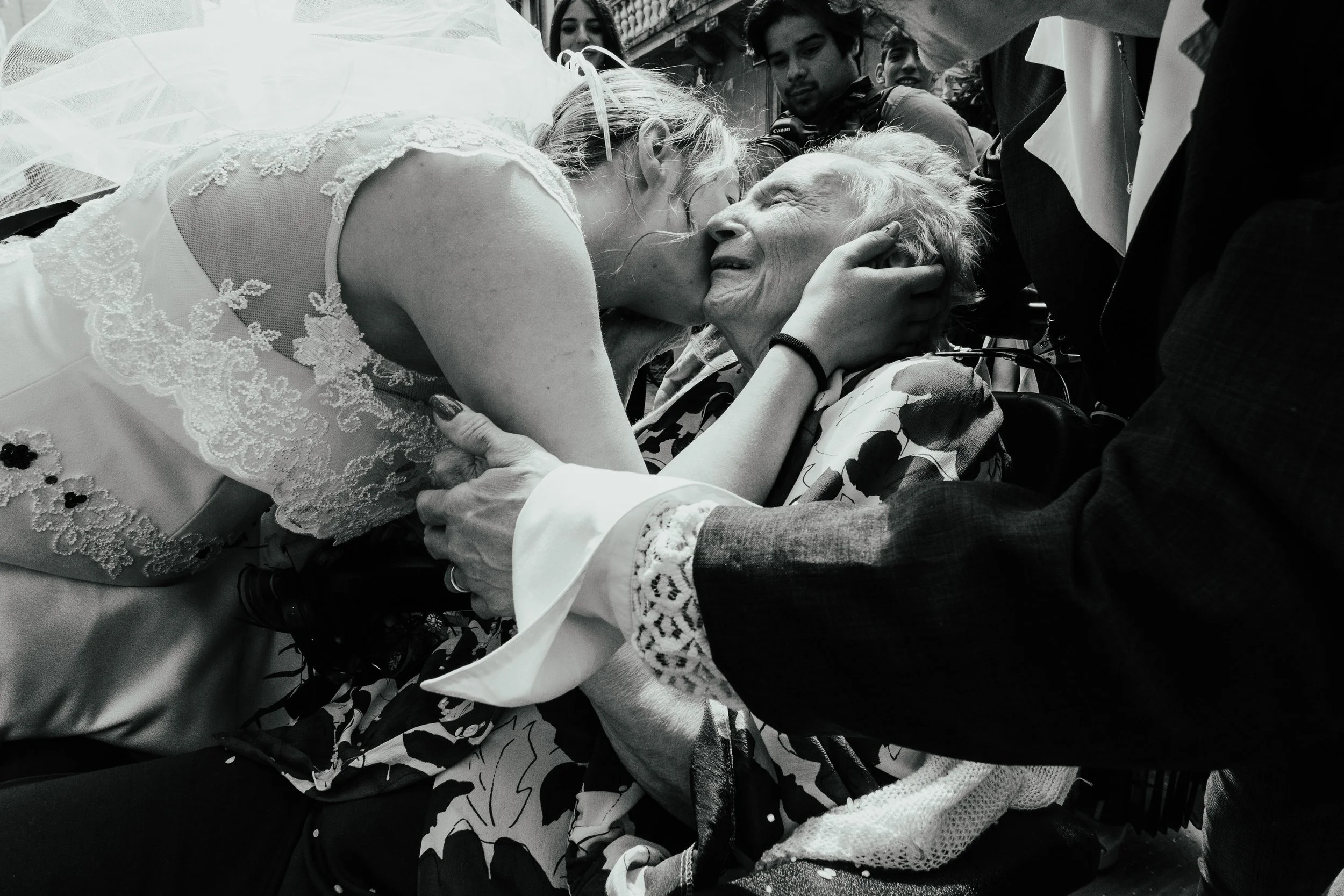 Una donna indossa un abito da matrimonio e bacia un'anziana in un momento emozionante, con altre persone intorno.