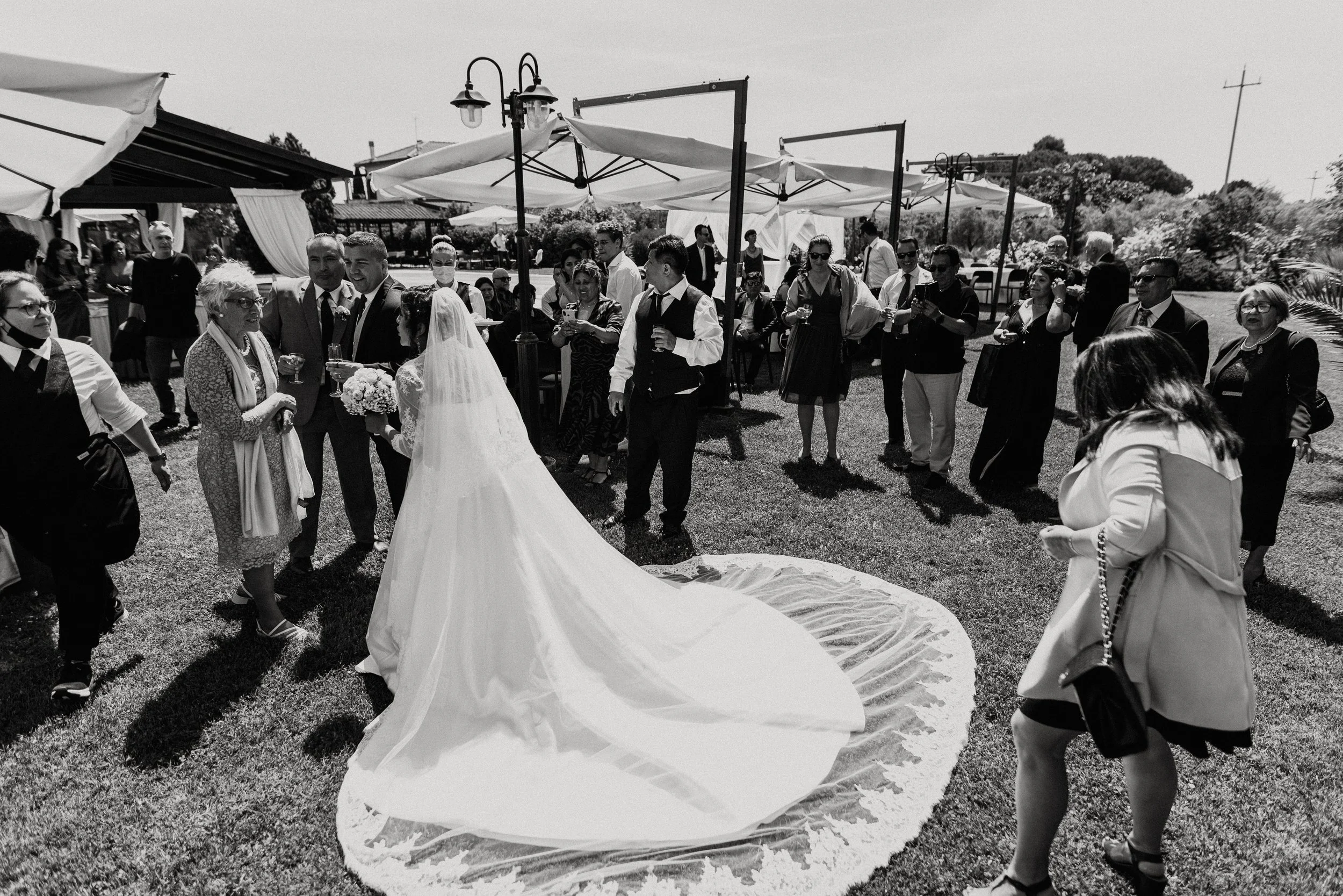 Celebrazione di matrimonio all'aperto con sposa, sposo e invitati che brindano, tutti vestiti eleganti, sotto ombrelloni e con alberi sullo sfondo.