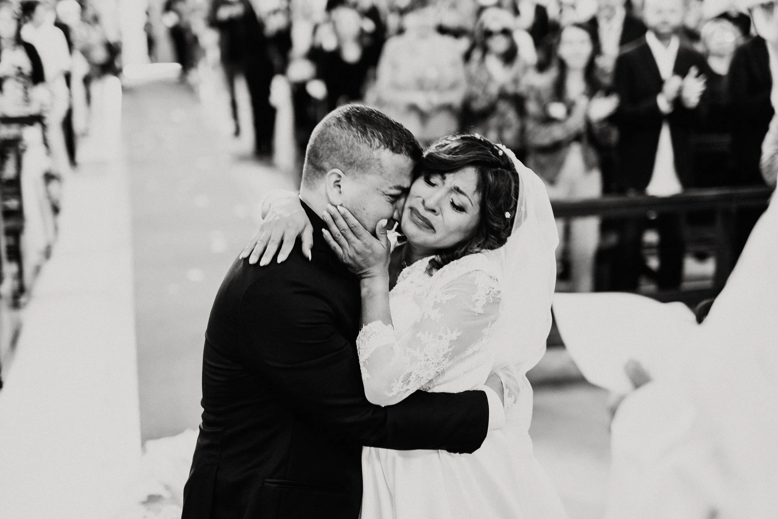 L'immagine mostra una coppia di sposi che si stanno abbracciando e piangendo durante il giorno del matrimonio in chiesa, con molti invitati sullo sfondo.