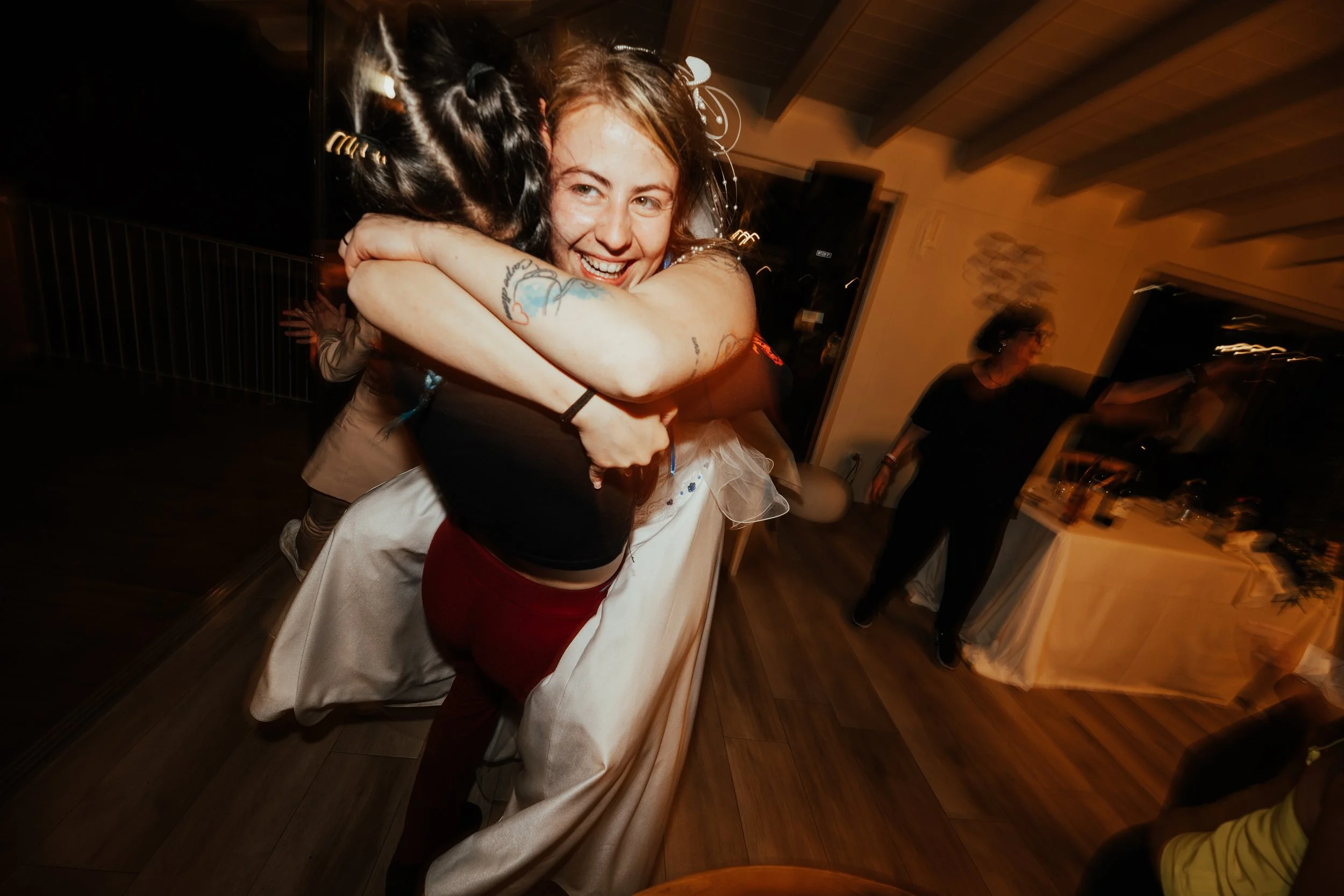 Due donne si abbracciano con gioia in un appartamento, sembra essere una celebrazione o una festa, con persone e decorazioni sullo sfondo