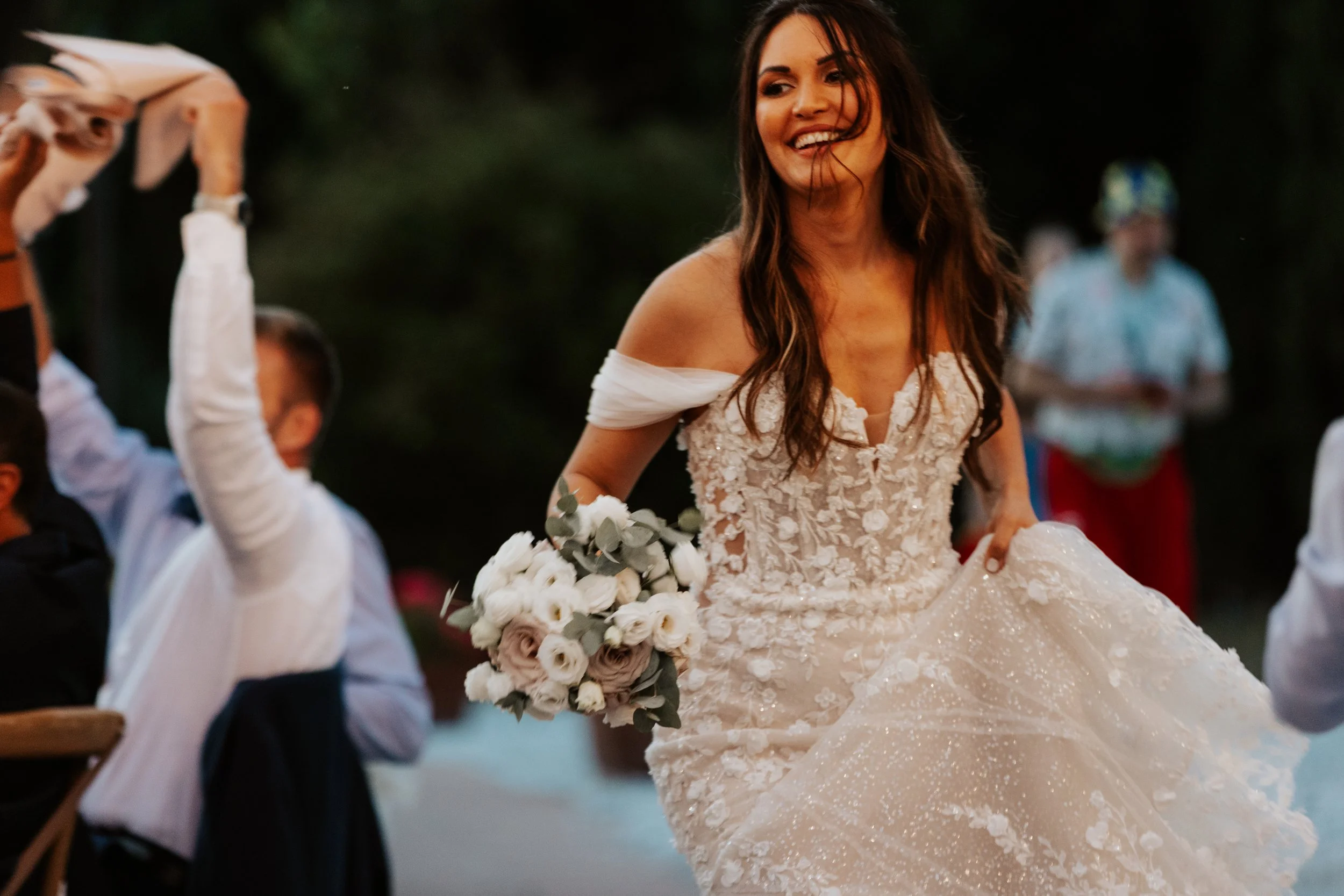 Una donna con abito da sposa che sorride camminando, tiene un bouquet di fiori bianchi e rosa, in un evento all'aperto.