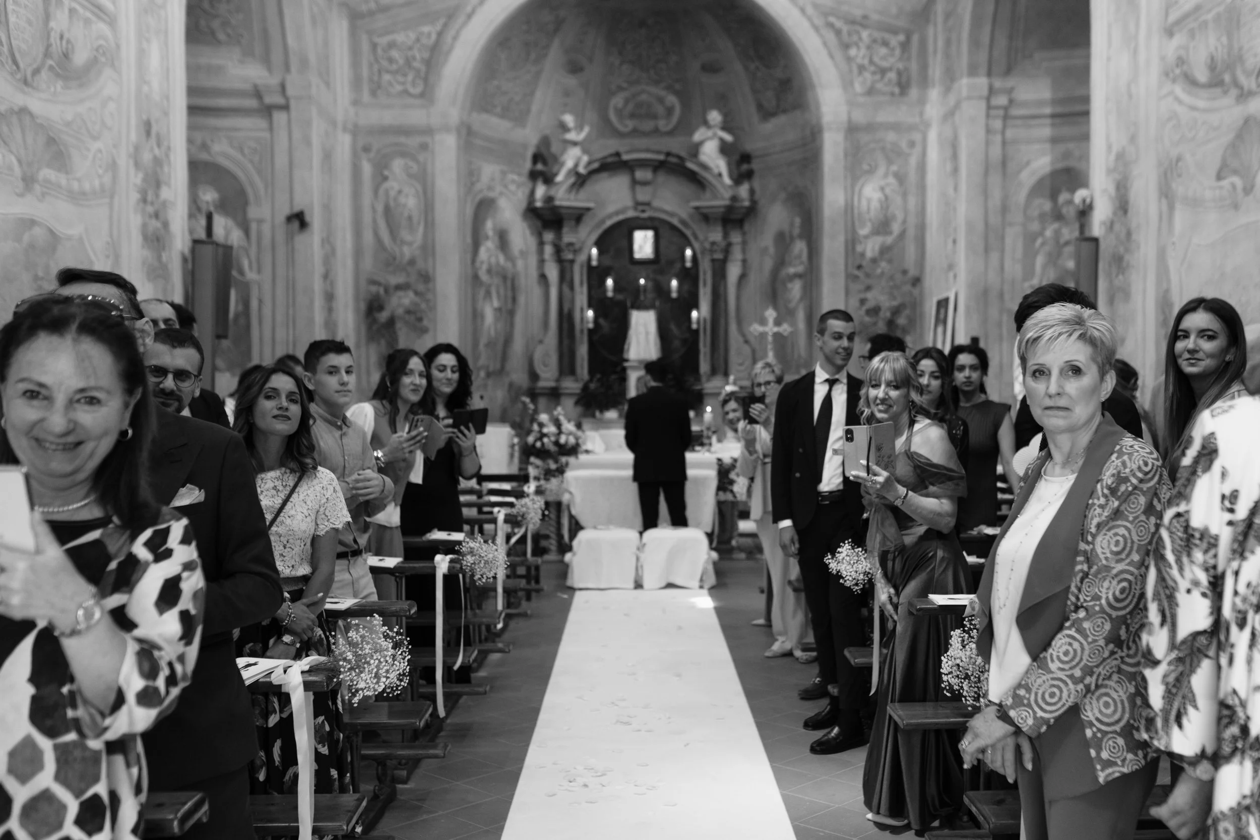 Una cerimonia di matrimonio in una chiesa con invitati che scattano foto e si riuniscono lungo il corridoio centrale.