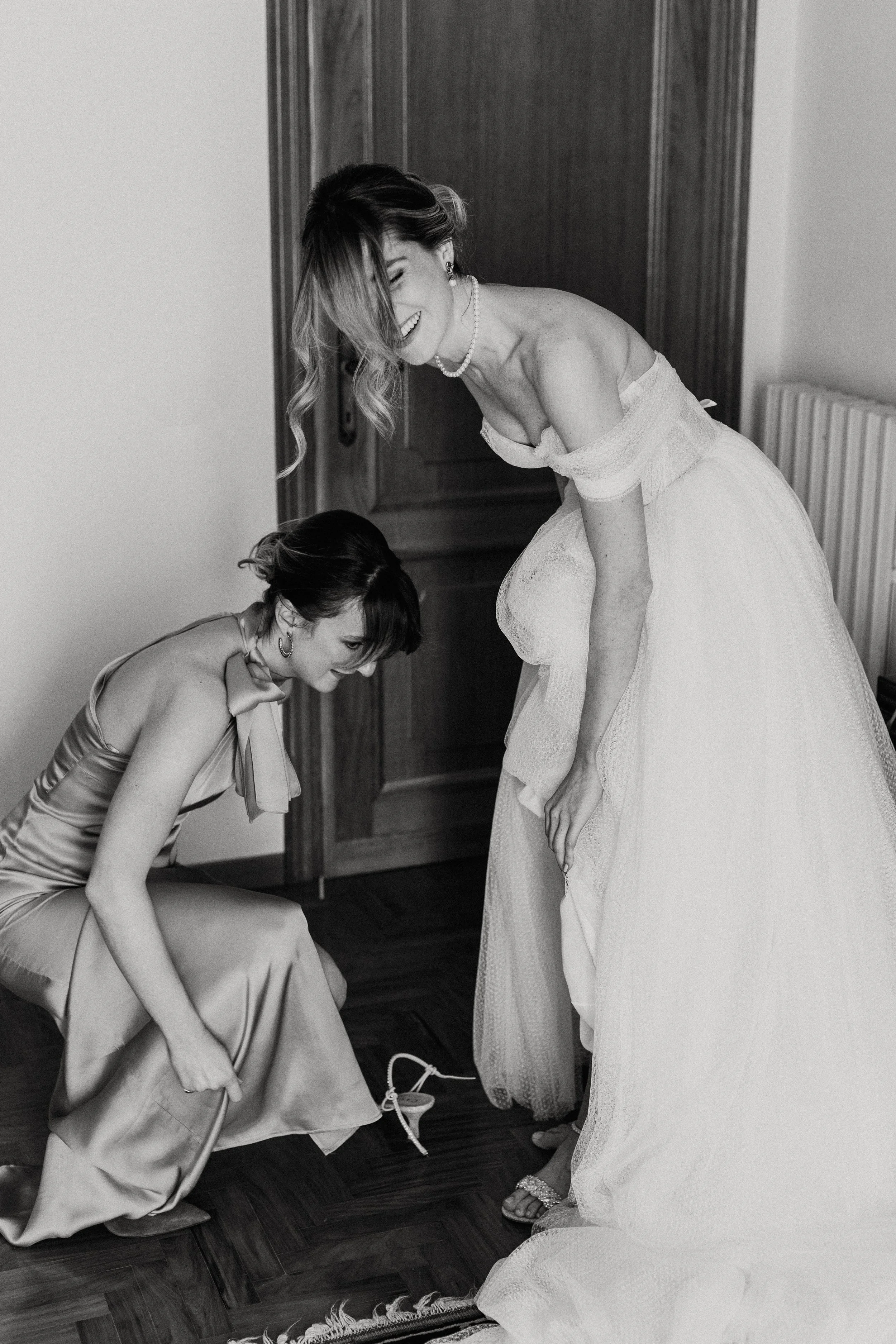 Donna in abito da sposa e un'altra donna in abito lungo si divertono ridendo e giocando tra di loro, in un ambiente interno.