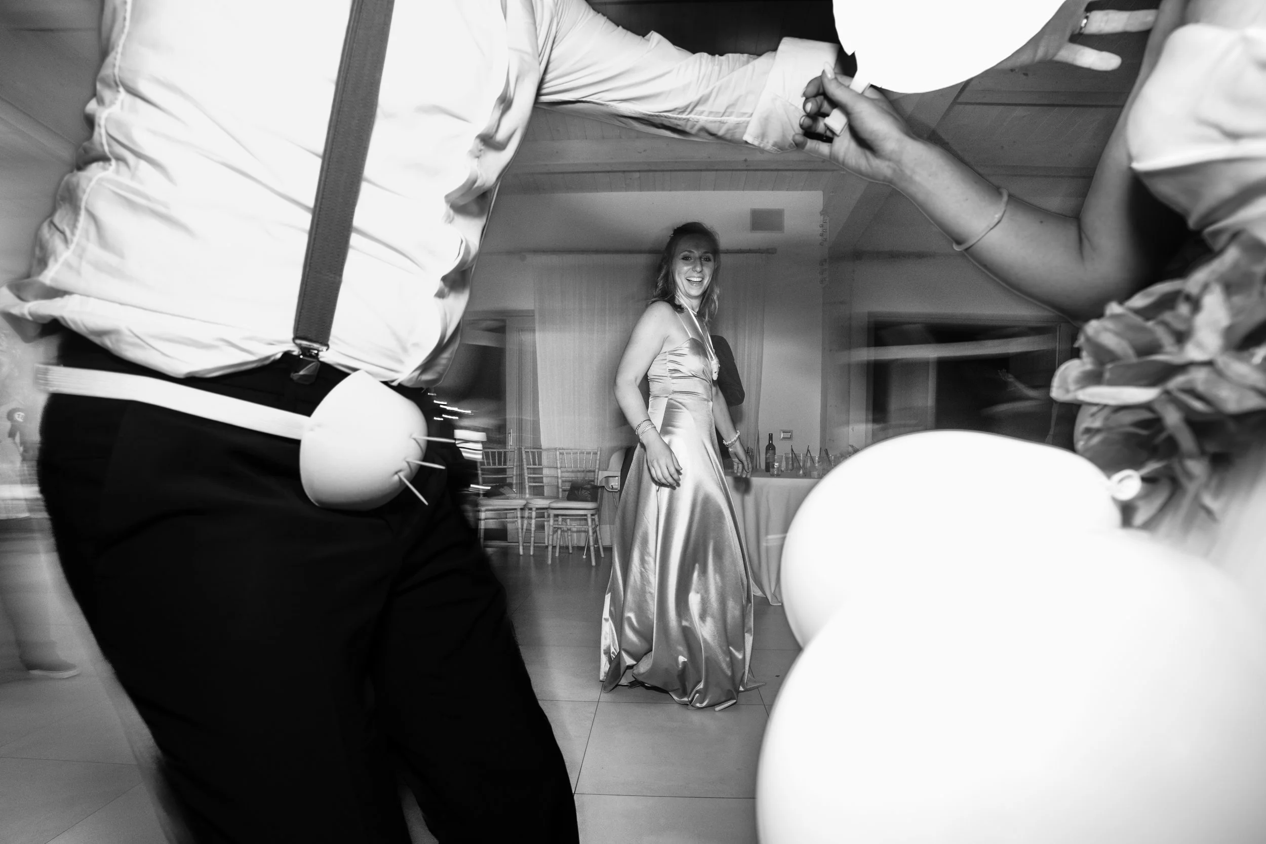 Una donna sorridente indossa un vestito lungo di seta e si trova al centro di una stanza, circondata da altre persone che tengono palloncini e si tengono per mano, in un'atmosfera di festa o matrimonio.