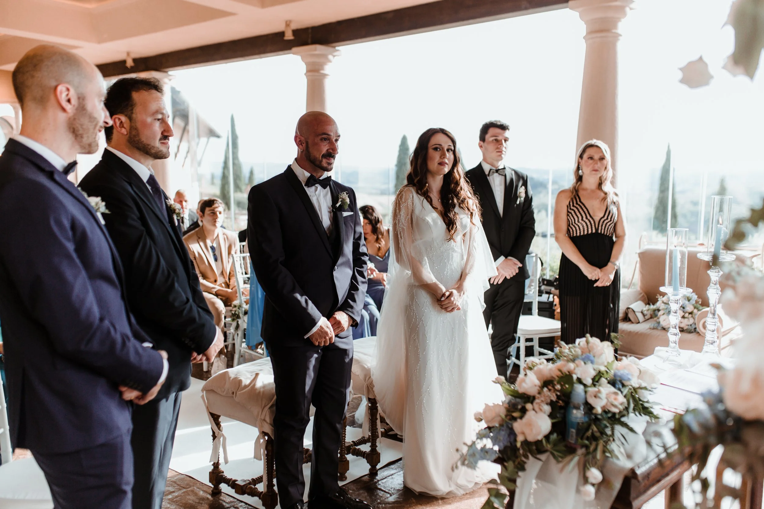 Coppia di sposi con testimoni e ospiti in un matrimonio in un luogo all'aperto con colonne e paesaggio di città sullo sfondo.