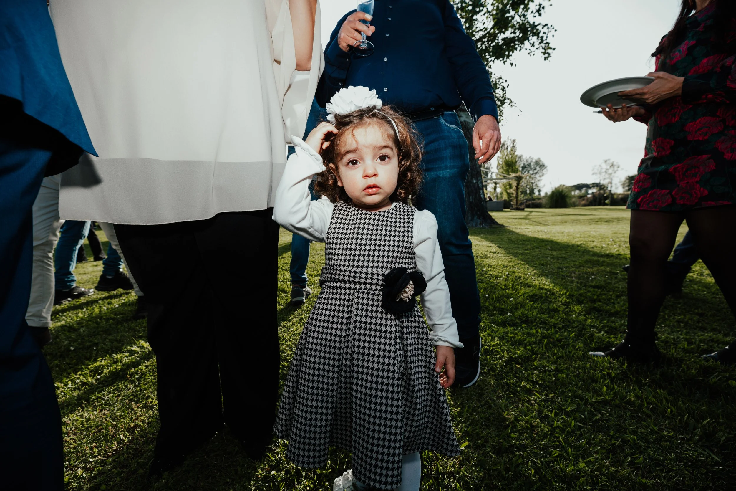 Bambina con vestito a scacchi nero e bianco e fiocco nero, che si trilla i capelli, circondata da adulti in un giardino, in una scena di festa o evento all'aperto.