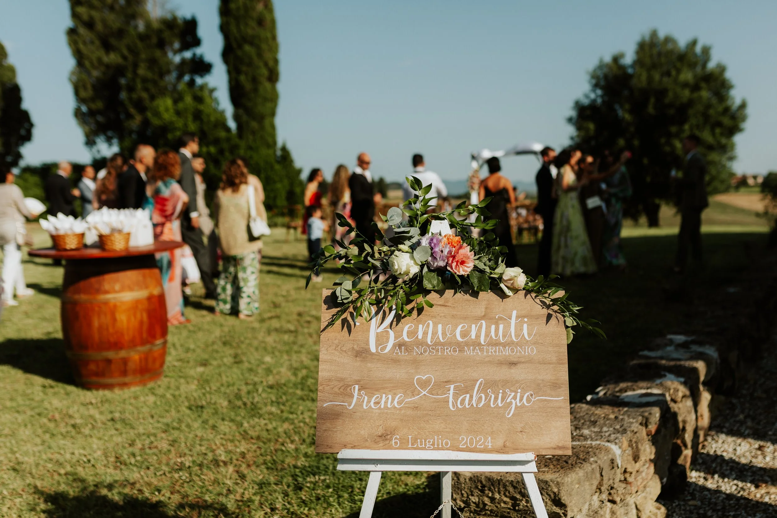 Cartello in legno con decorazione floreale che dice 'Benvenuti al nostro matrimonio, Irene e Fabrizio, 6 Luglio 2024' in un matrimonio all'aperto con persone riunite in sfondo. Agriturismo Cà Lo Spelli. Del Carlo Catering