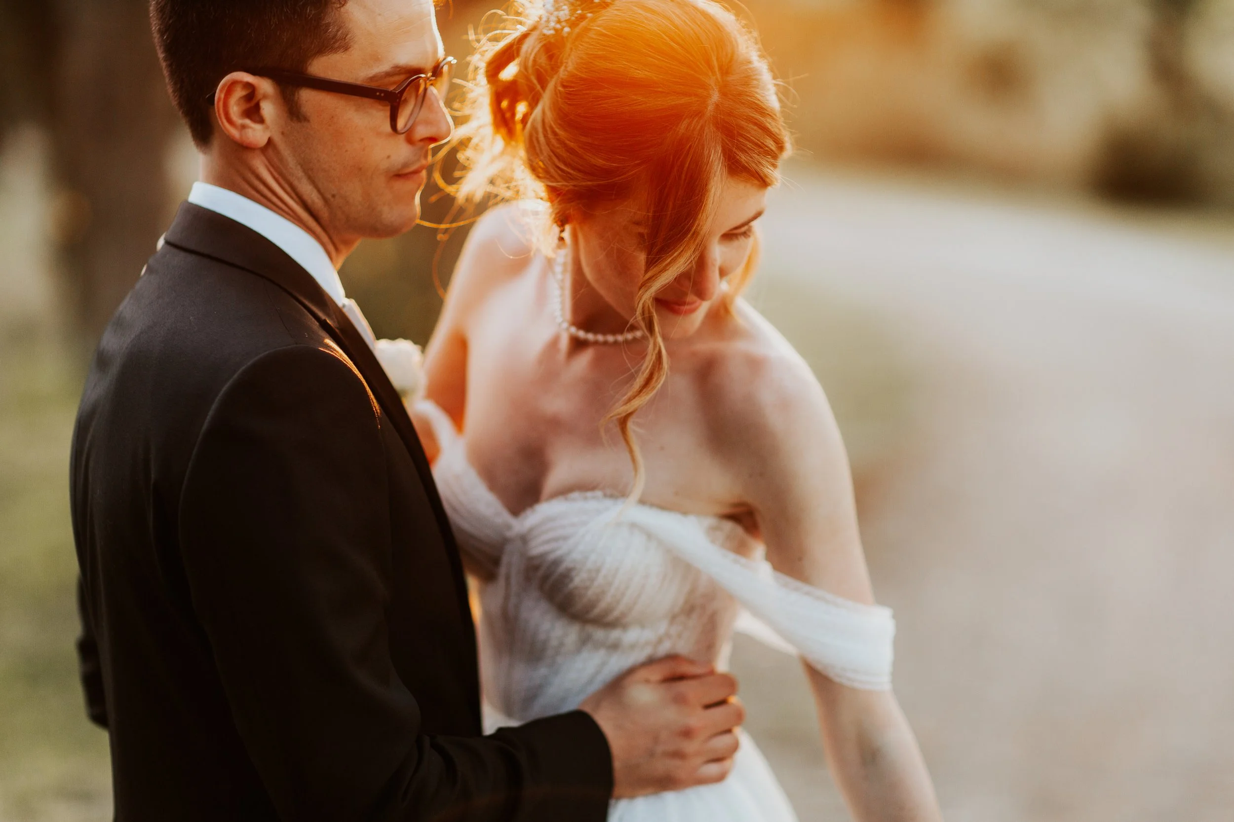 Coppia di sposi al tramonto, uomo in completo nero e donna in vestito bianco, in un ambiente naturale.