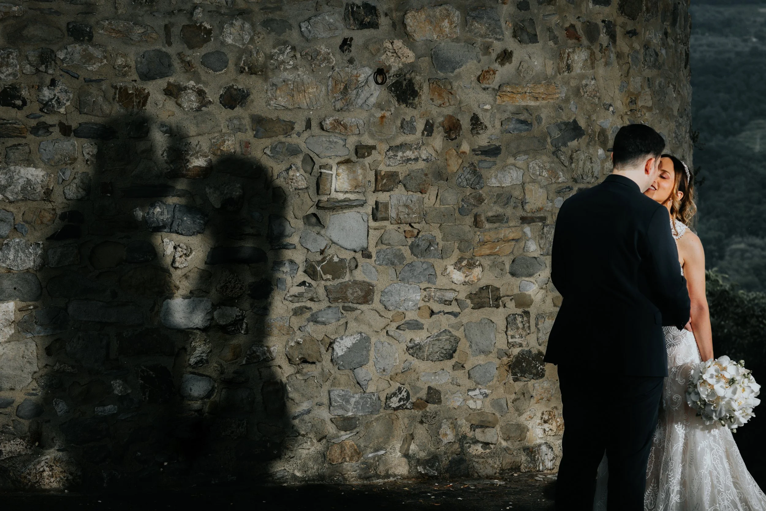 Una coppia in abiti da matrimonio si bacia davanti ad un muro di pietra, con l'ombra della donna proiettata sulla parete.