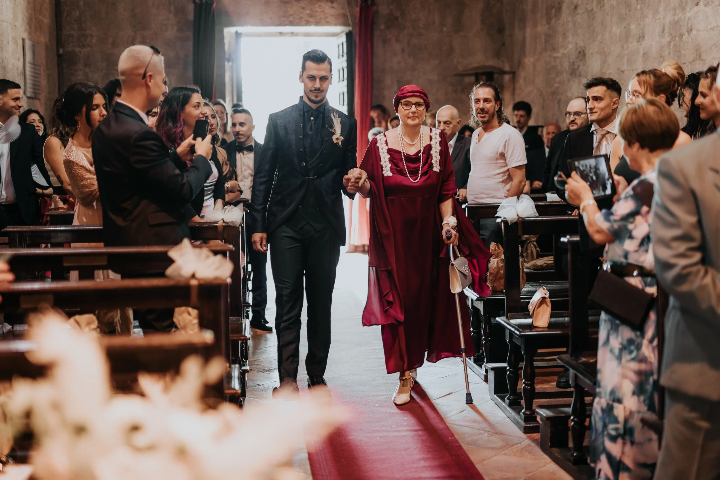 Una donna anziana in abito rosso con cravatta bianca, con bastone e borsa, accompagna un giovane uomo in abito scuro, davanti a una chiesa durante un matrimonio. Circondati da amici e parenti che li fotografano e sorridono.
