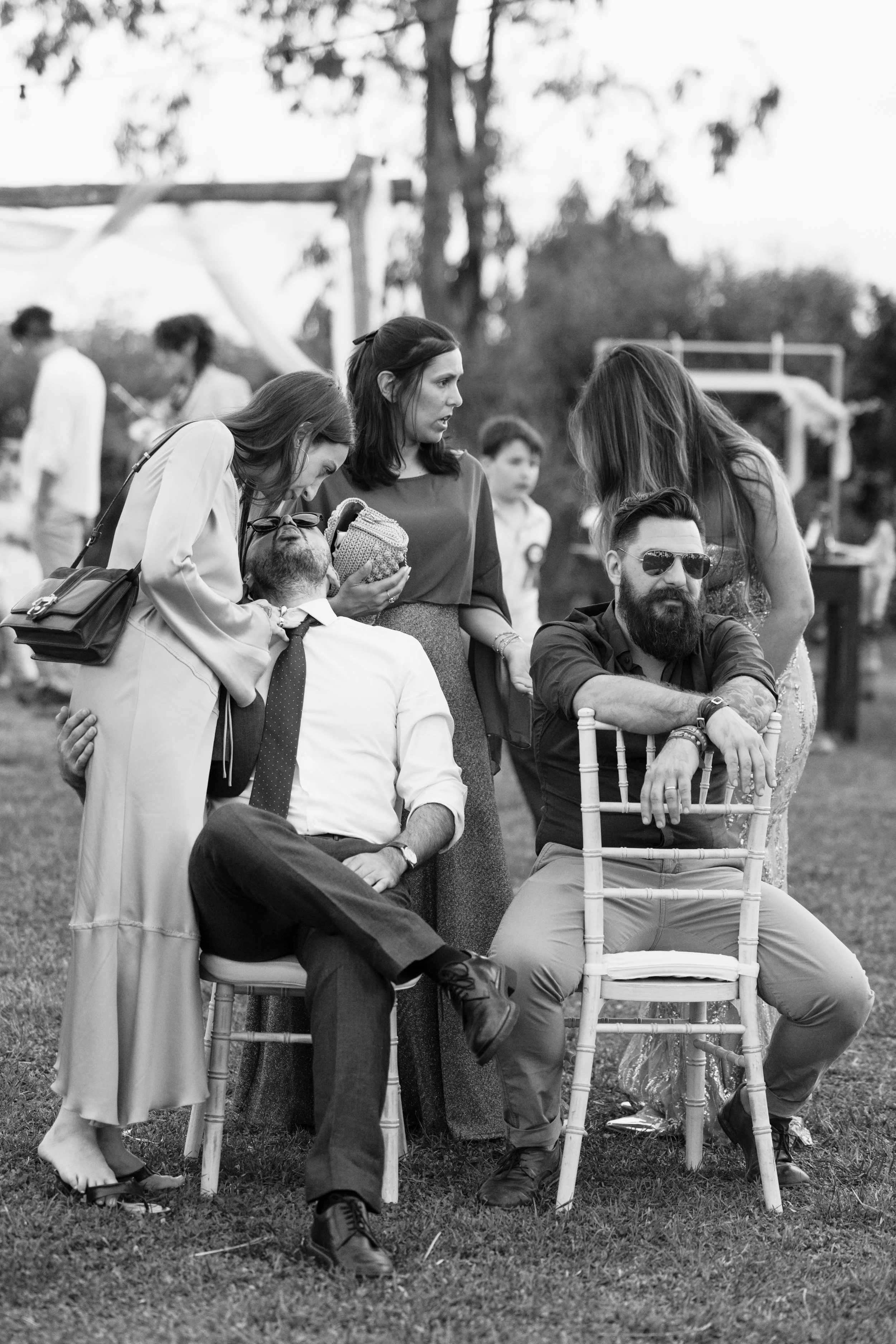 Un gruppo di persone, tra cui due uomini seduti su sedie e tre donne, sono in un cortile durante un evento sociale all'aperto. Sono in un contesto festoso, forse un matrimonio o una festa.