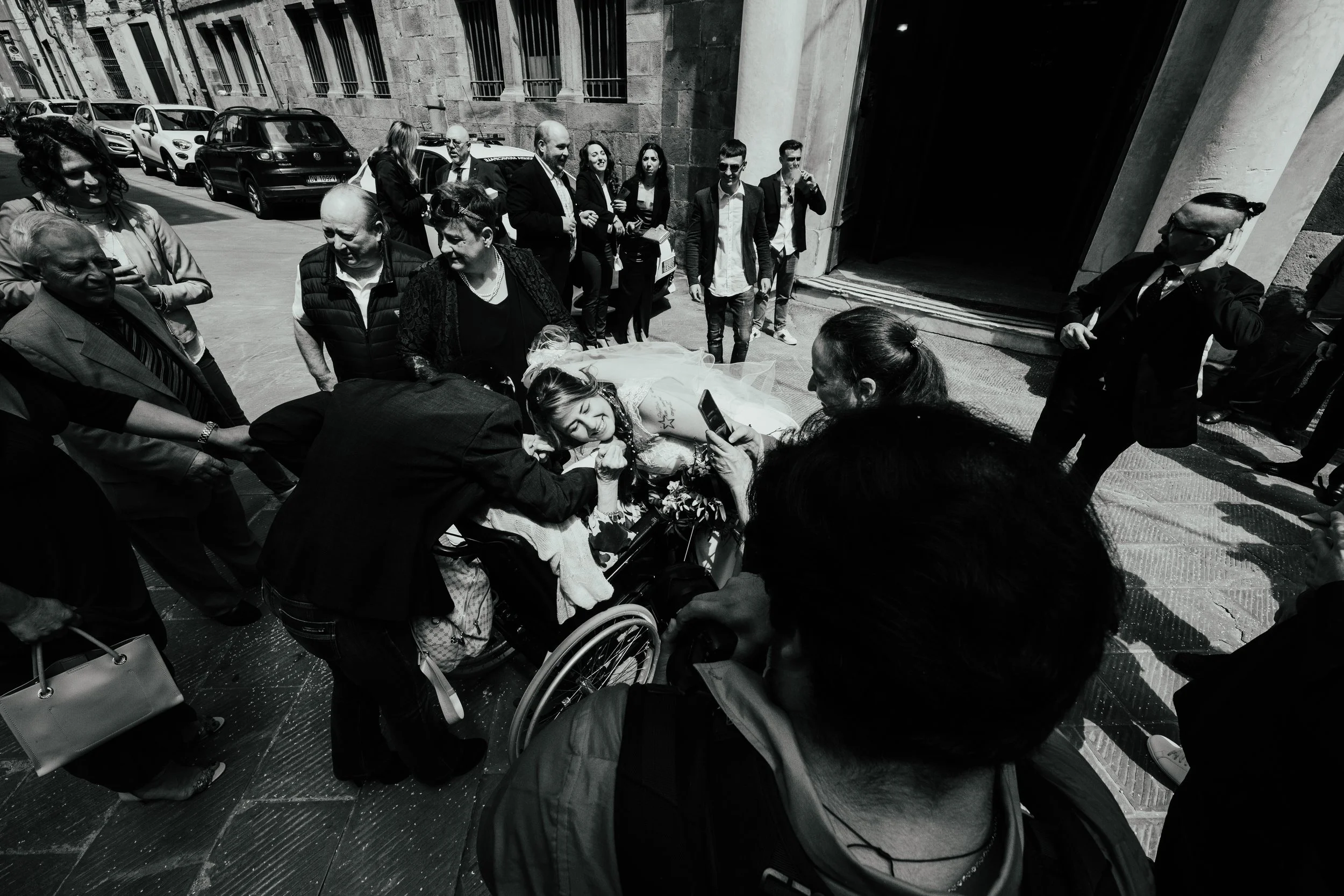 Una donna in sedia a rotelle riceve supporto e abbracci da persone circostanti, mentre alcune scattano foto, in una strada urbana.