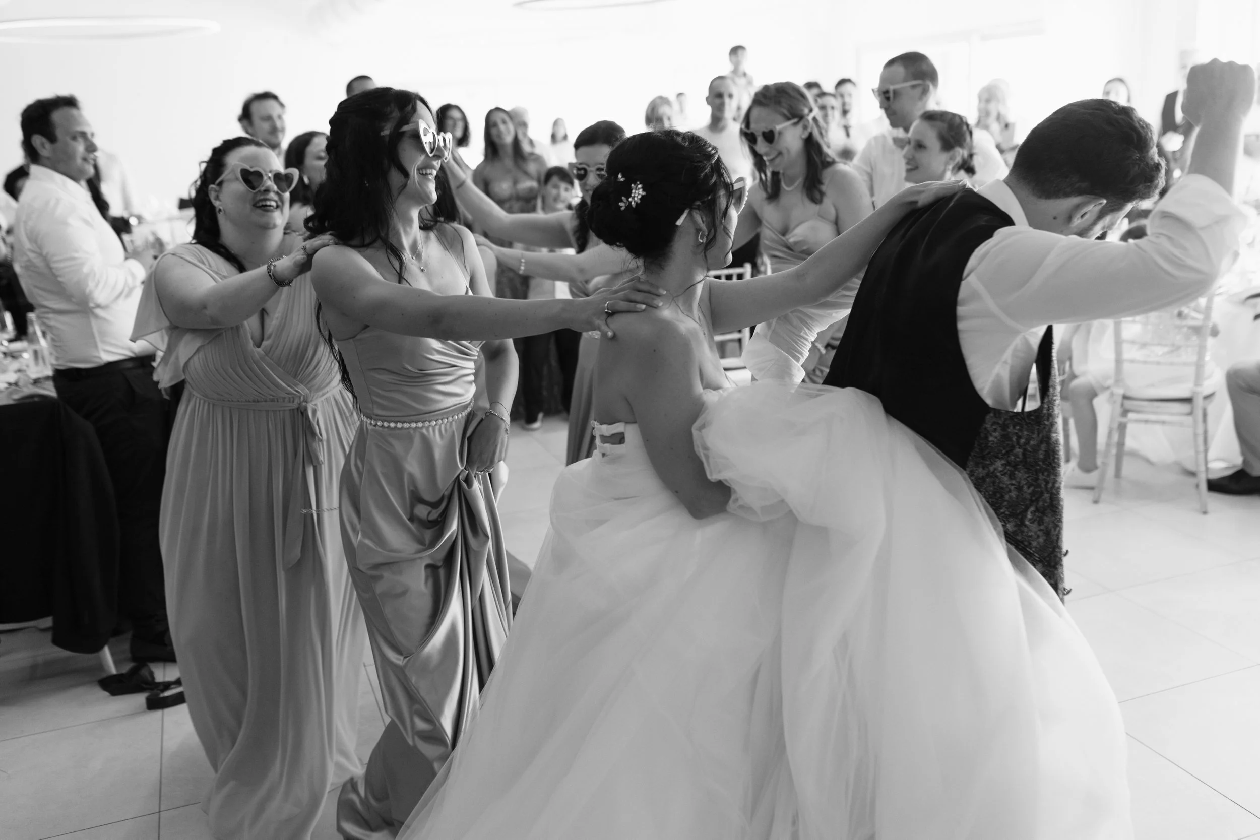 Foto di una festa di matrimonio con persone che ballano in cerchio, alcune indossano occhiali divertenti, alcune sono sedute e altre in piedi, in un ambiente luminoso.