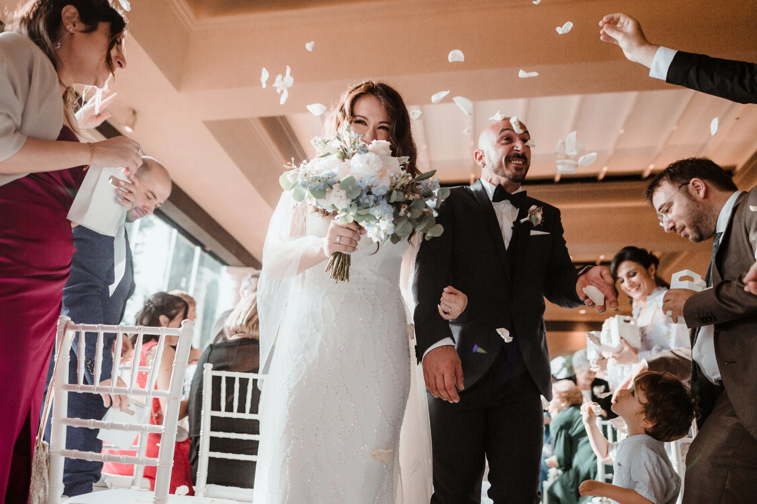 Una coppia di sposi felici durante il matrimonio, circondata da amici e familiari che lanciano petali di fiore, con l'atmosfera gioiosa e celebrativa dell'evento.