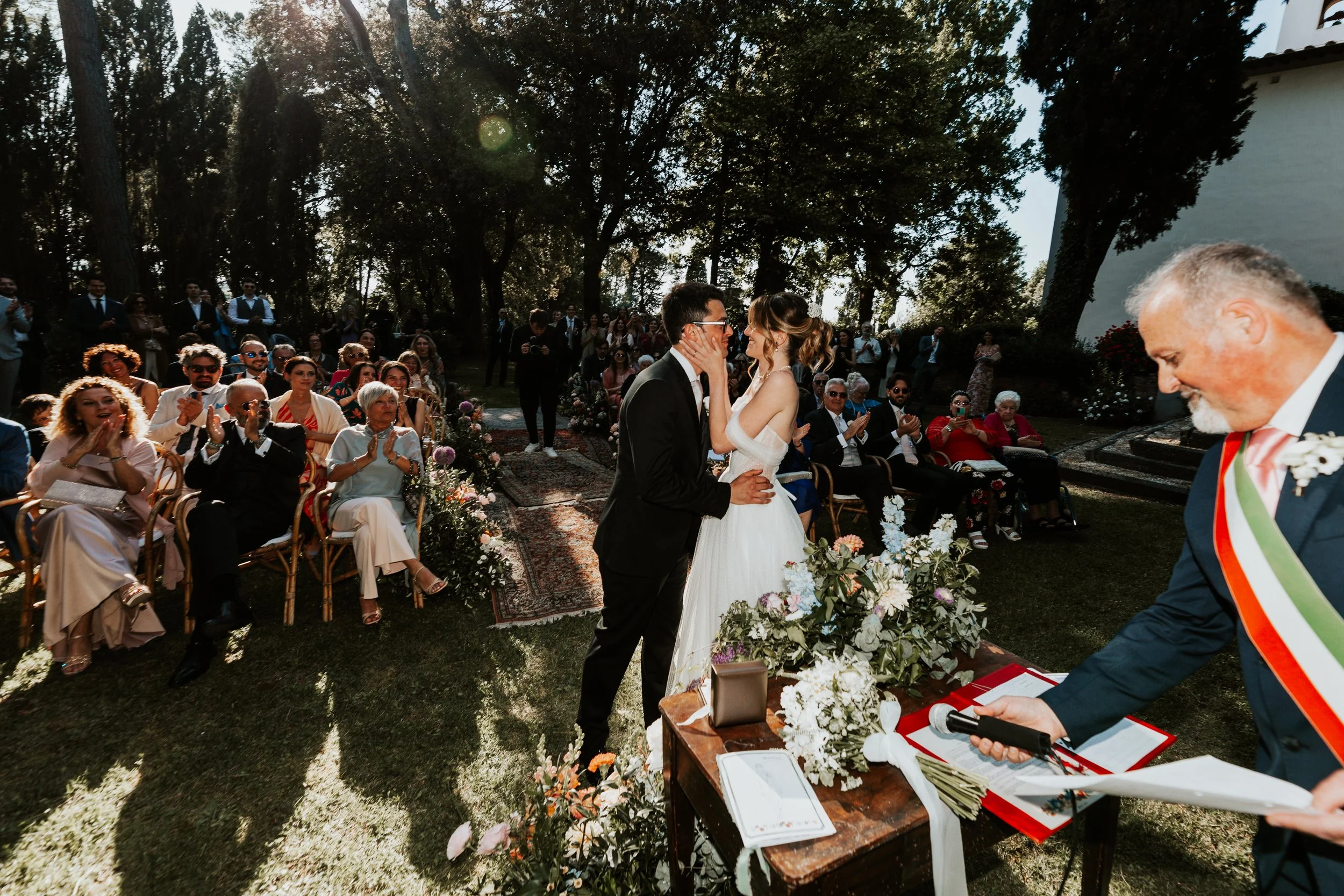 Partecipanti a un matrimonio all'aperto, con il sindaco che legge i voti, coppia di sposi al centro, amici e parenti che applaudono, ambiente naturale con alberi e decorazioni floreali.